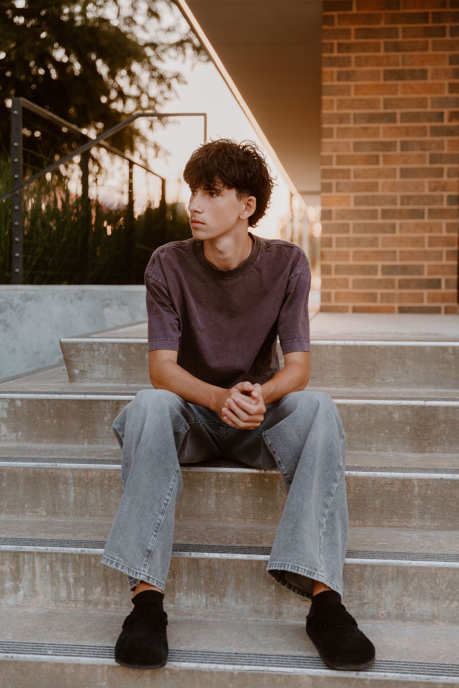 Person sitting on concrete steps wearing a dark t-shirt and jeans at sunset with brick wall in background.