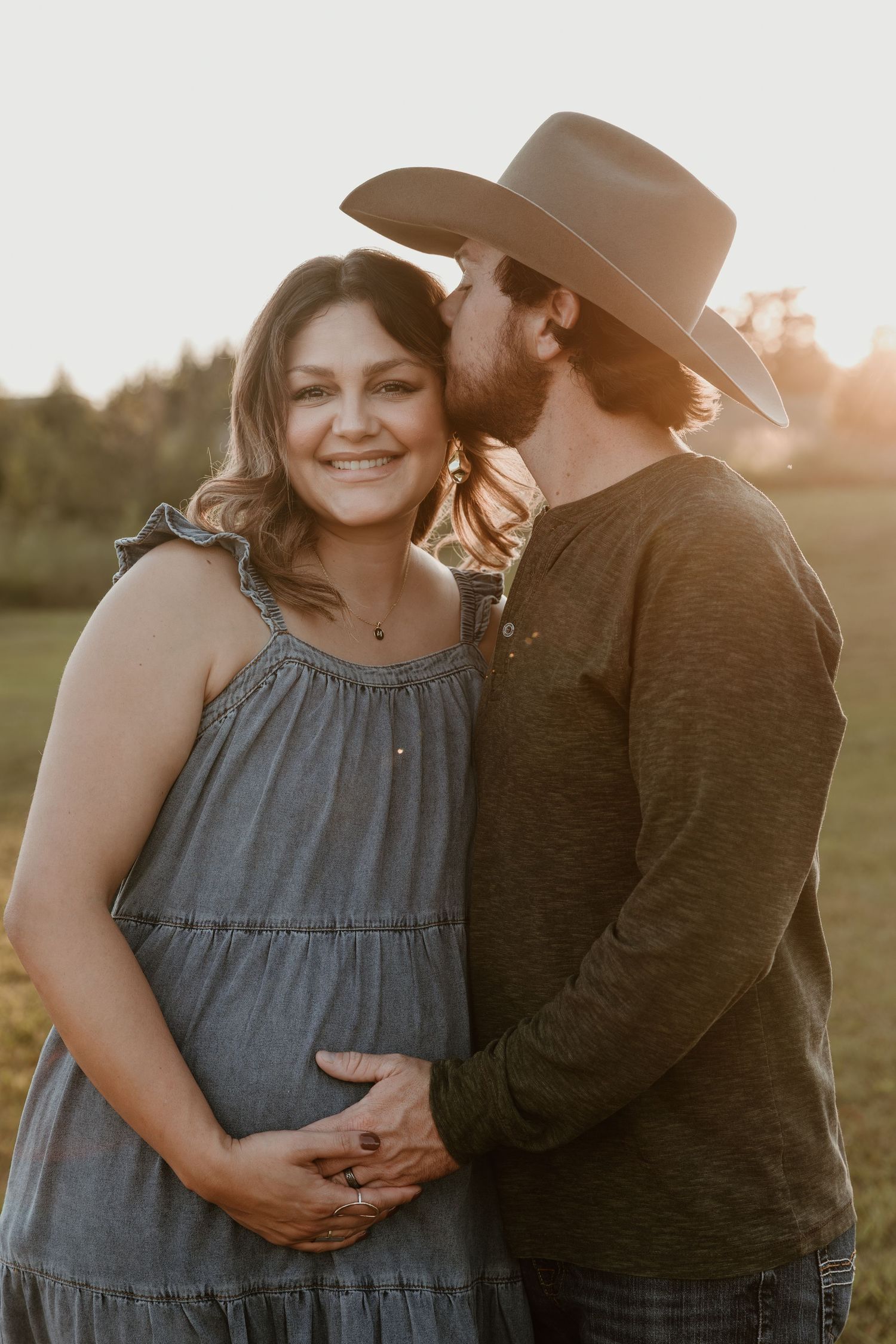 A romantic sunset maternity photo session showing a couple embracing in a field wearing western style clothing.