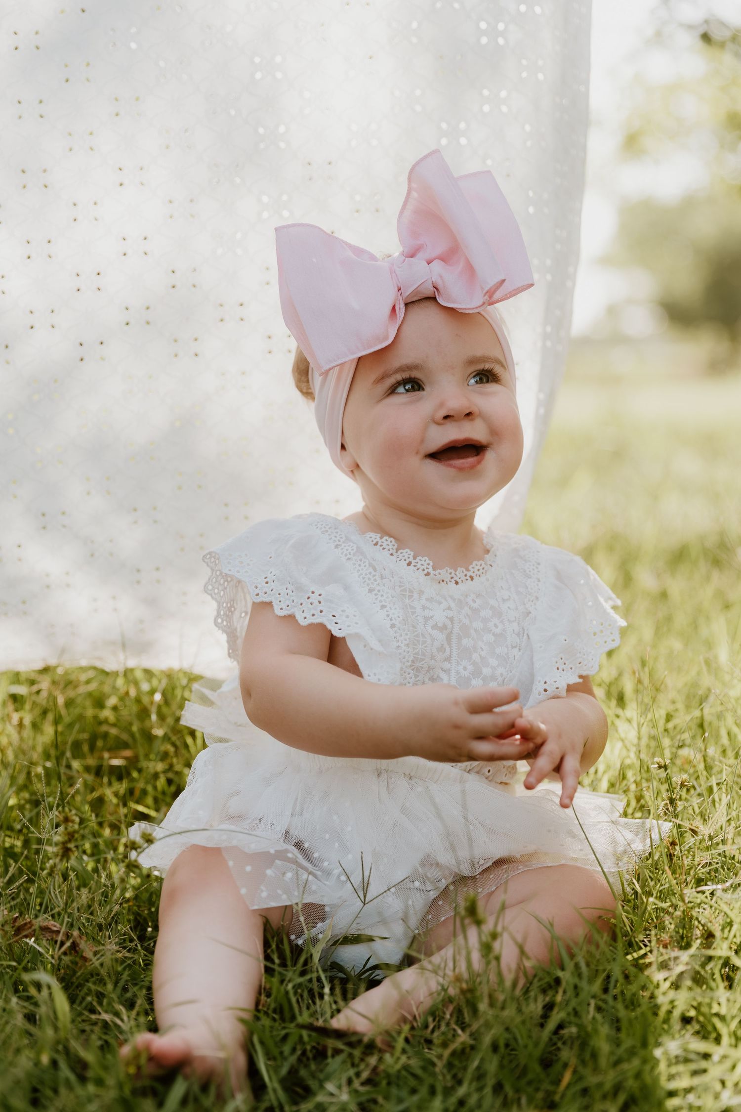 Adorable baby girl in white lace romper and pink bow headband sitting on green grass outdoors.