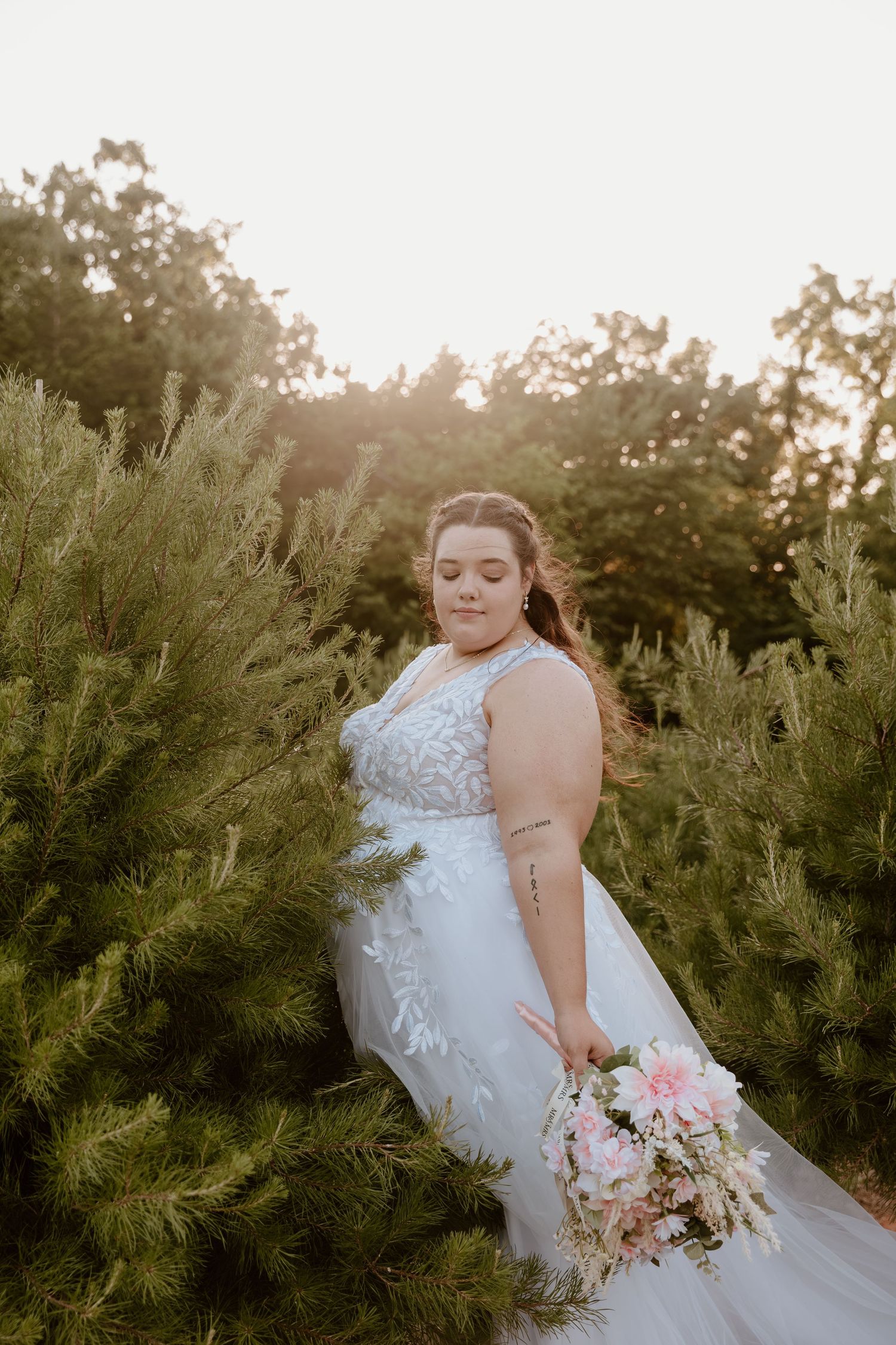 Bride in lace wedding gown holds pink bouquet among evergreen trees in golden hour light.