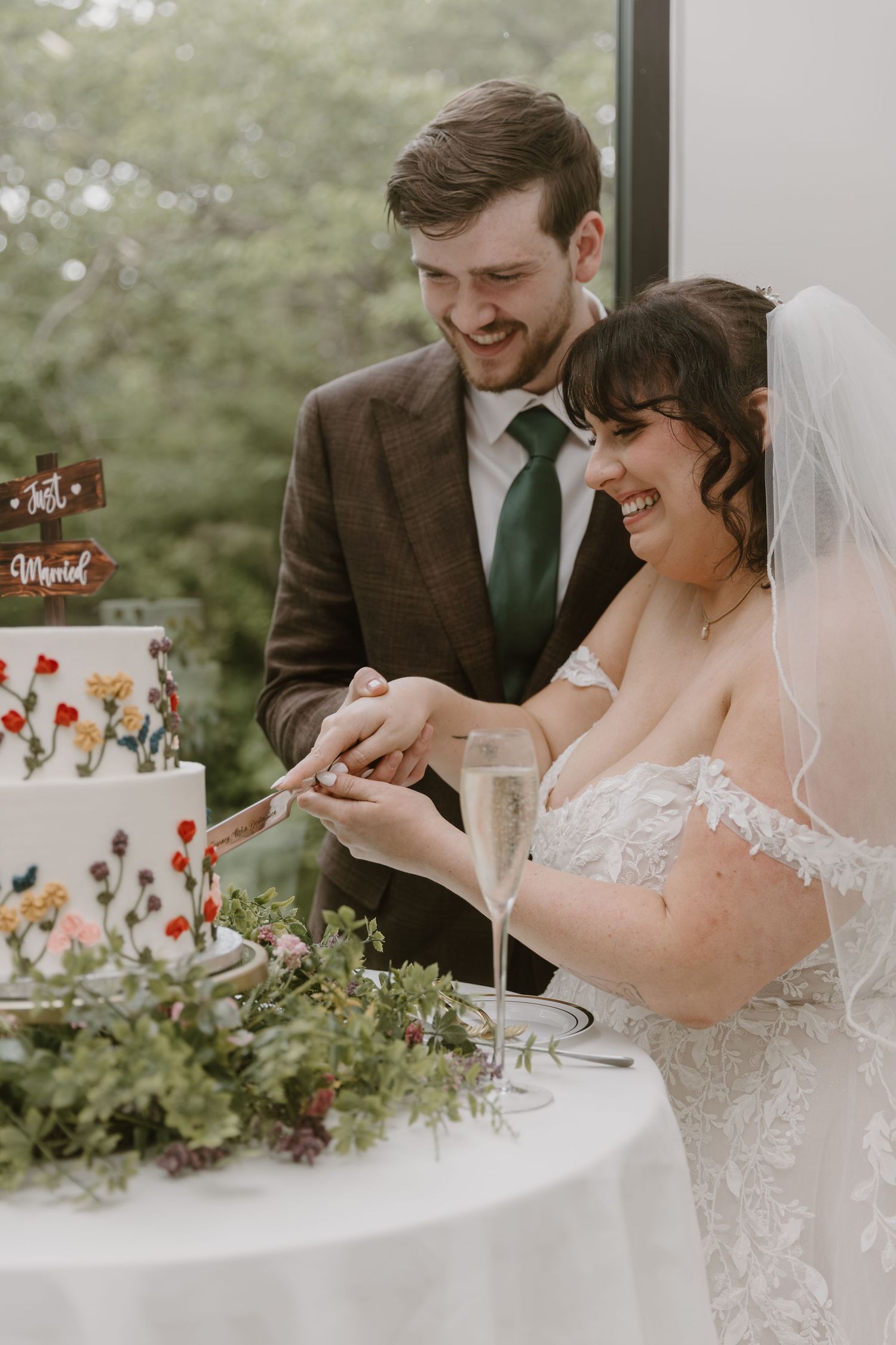 Bride and groom smiling while cutting their floral wedding cake, with champagne glasses nearby.