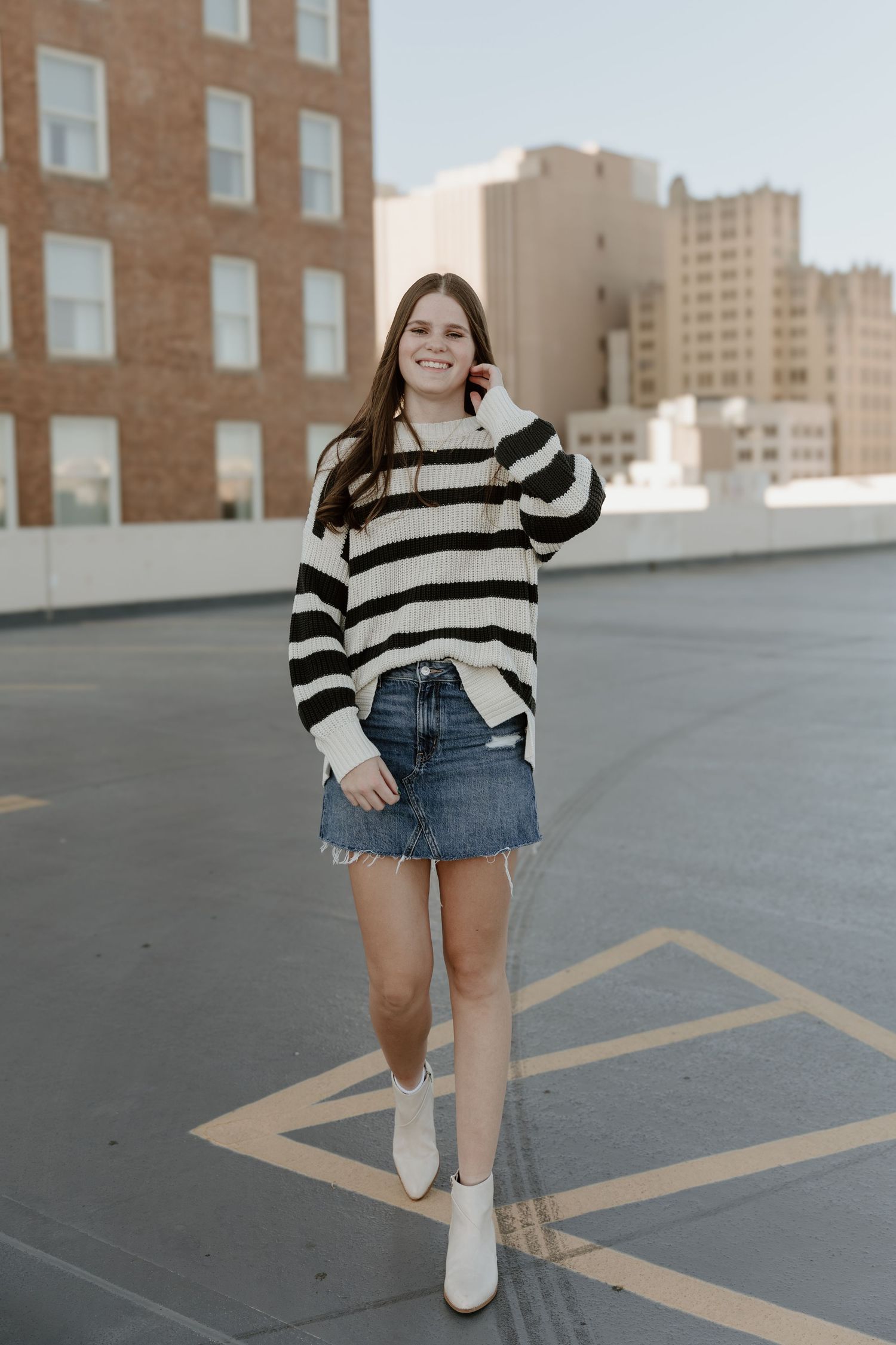 A person in a striped sweater and denim shorts poses on a rooftop parking lot with city buildings in the background.