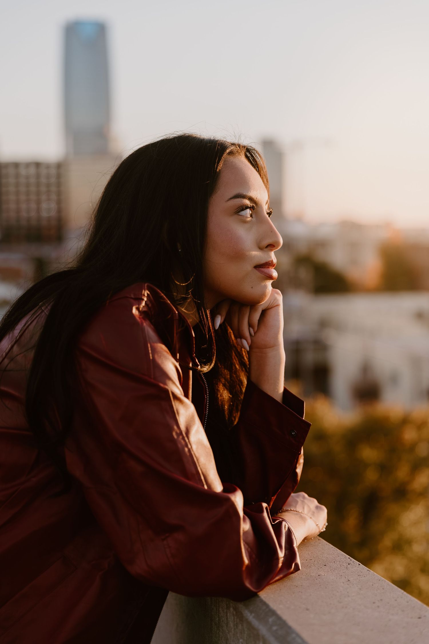 A person in brown clothing gazes thoughtfully at sunset against an urban backdrop with warm golden lighting.