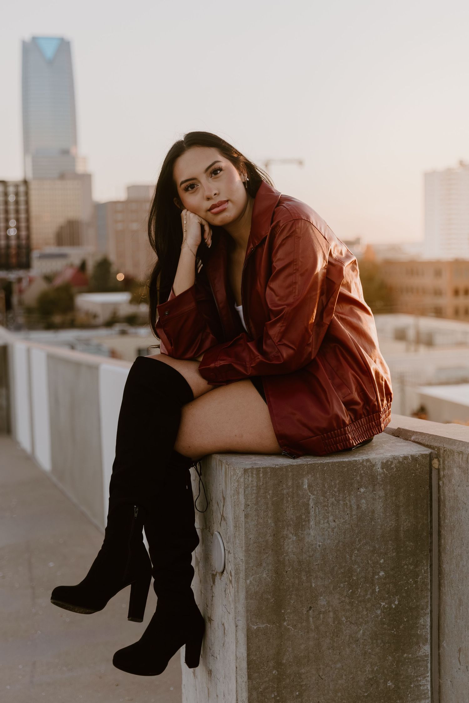 A person in a burgundy jacket and knee-high boots sits on a concrete ledge with a cityscape backdrop at sunset.