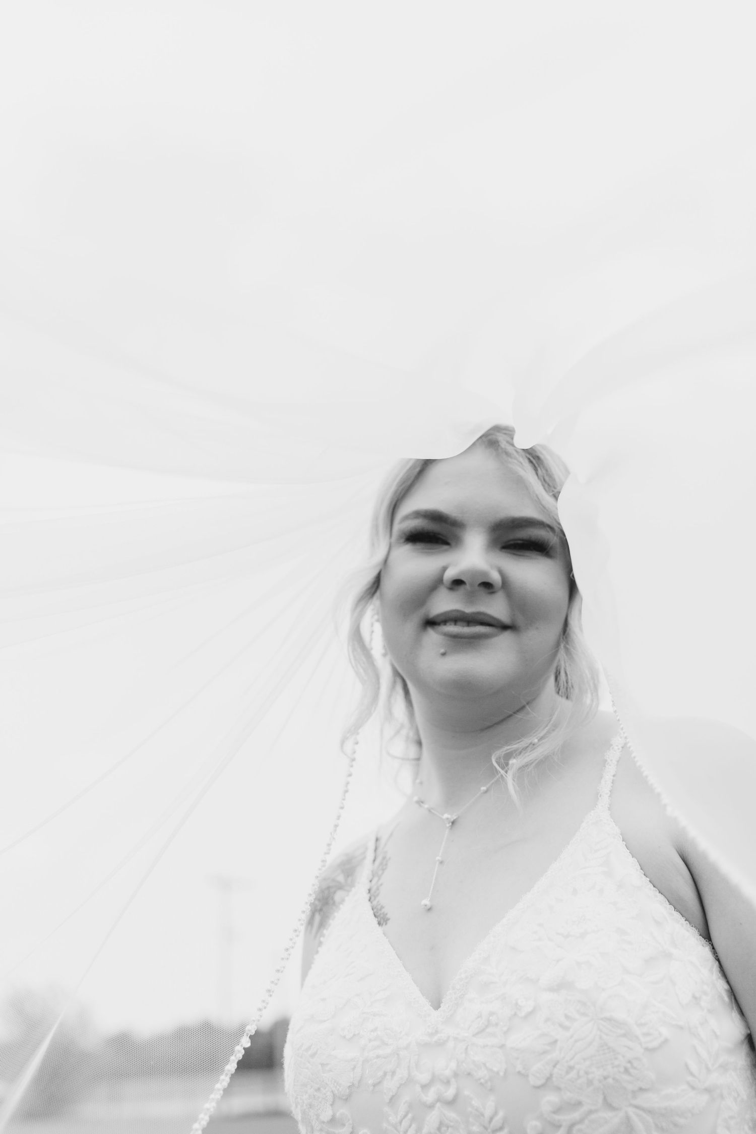 A bride in a white lace wedding dress and veil smiles while looking down at the camera in black and white.