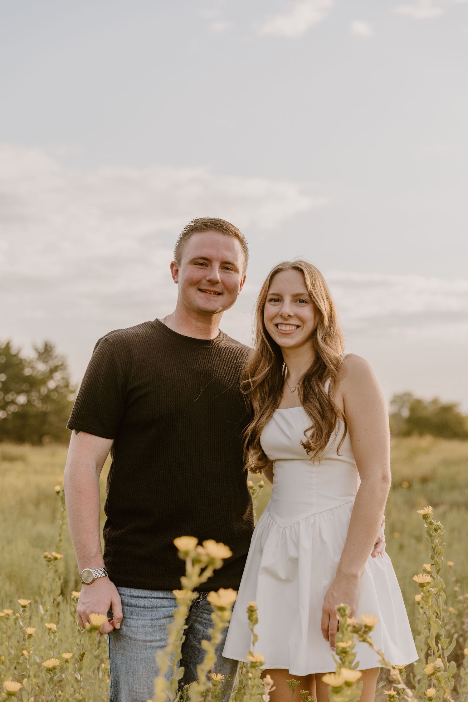 A couple stands together in a wildflower field during golden hour, wearing casual summer attire.