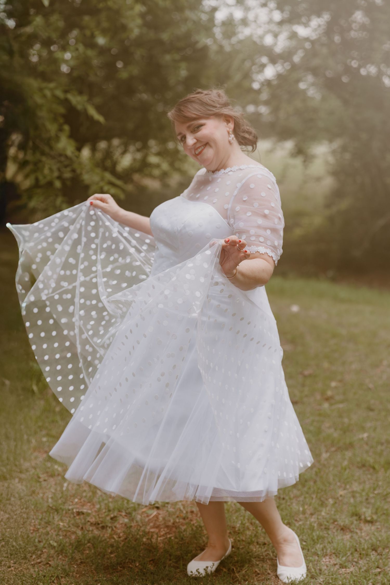 Smiling person in white lace tea-length wedding dress spins outdoors while holding polka dot fabric.