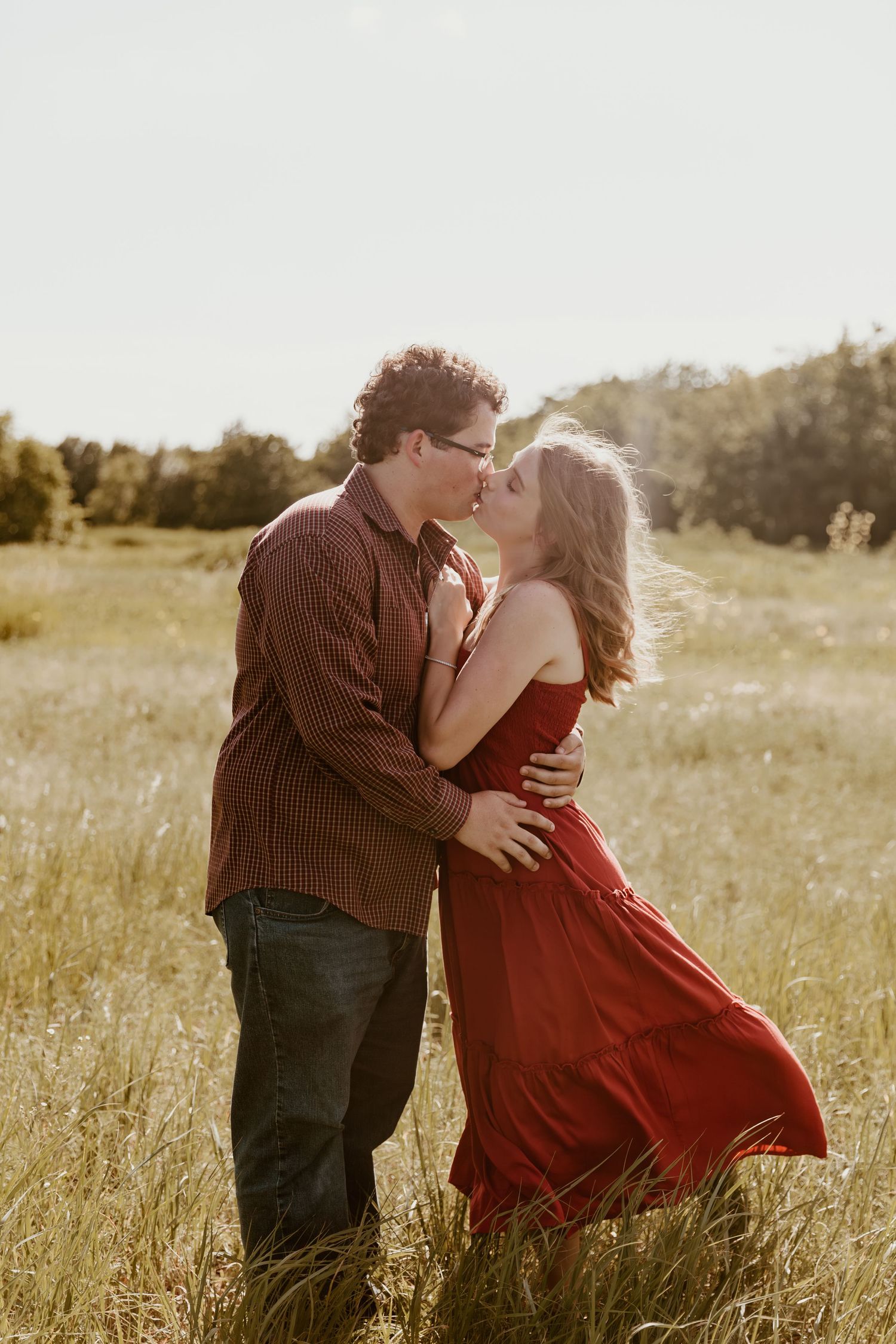 A romantic couple embraces in a golden sunlit field as her red dress flows in the breeze during sunset.