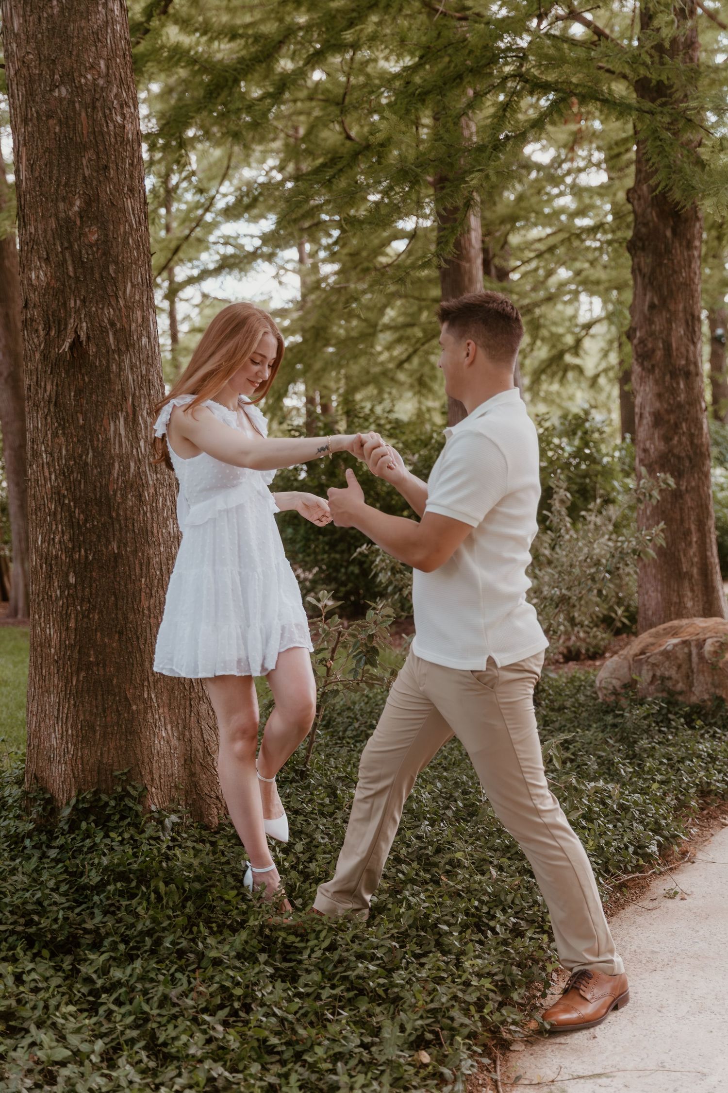 A couple in coordinated white and beige outfits share a romantic moment dancing together on a wooded path.