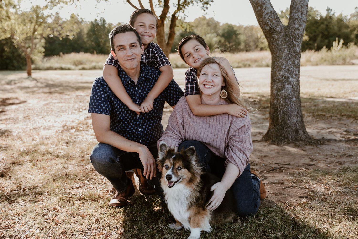 A couple and their Papillon dog pose together for outdoor family photos in a sun-drenched park setting.