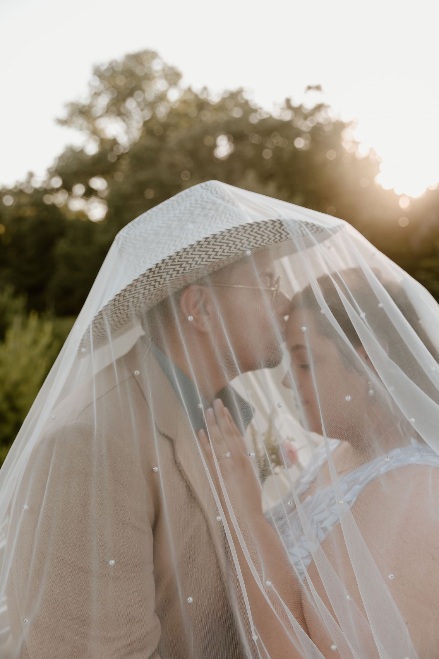 Romantic bride and groom kissing under bridal veil in golden sunset outdoor wedding photo.