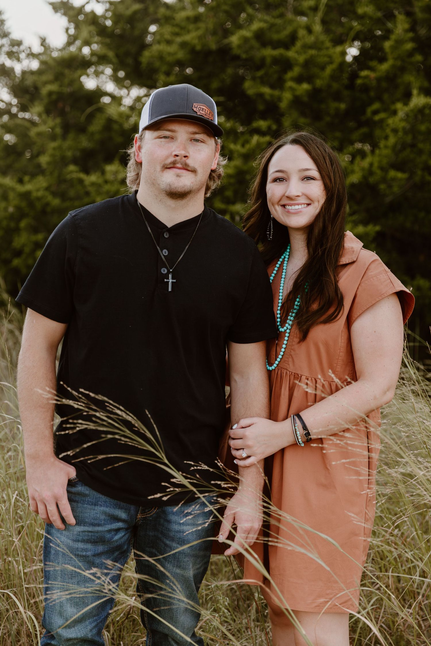 A couple poses together in tall grass during an outdoor autumn photo session, wearing casual attire.
