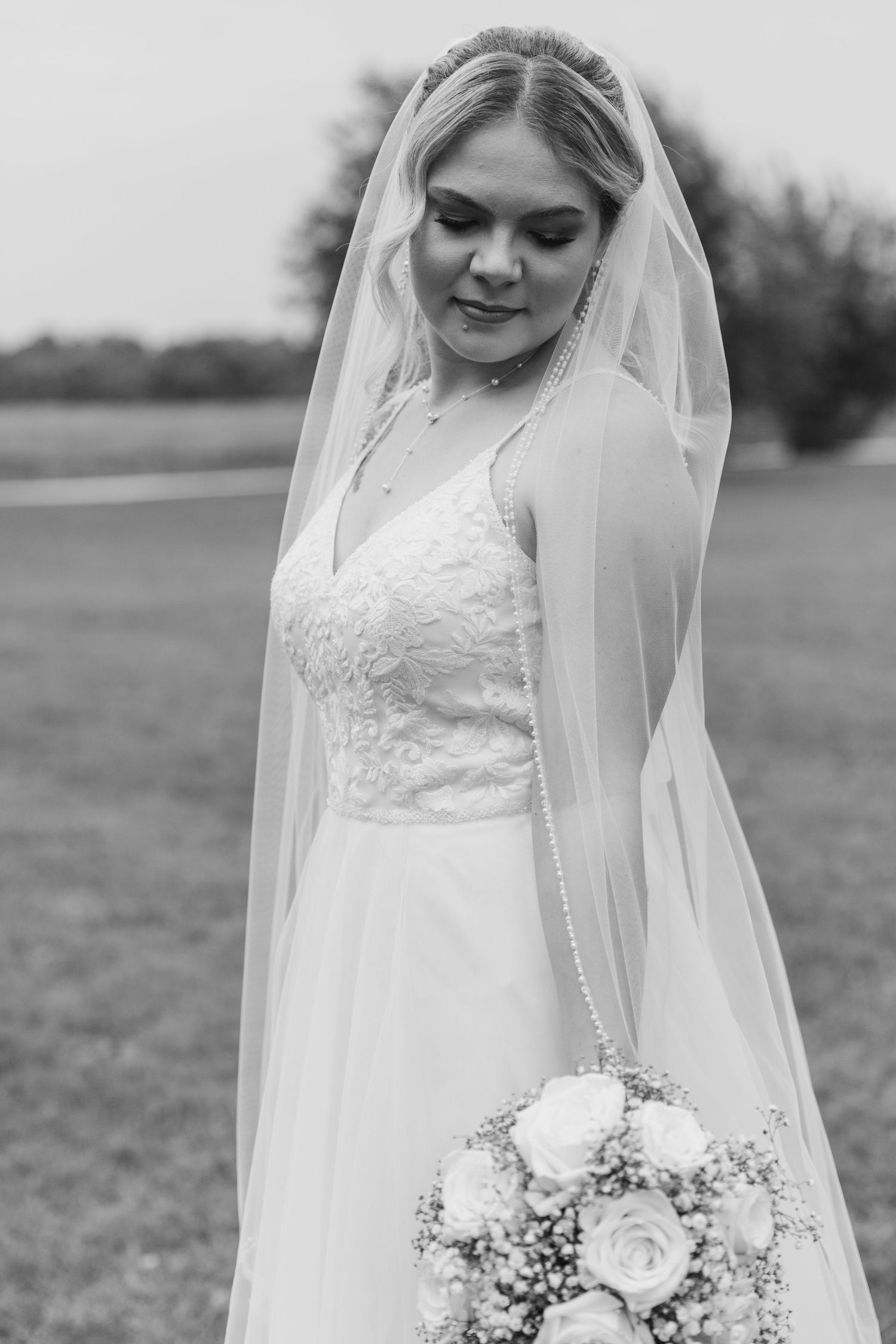 Black and white photo of bride in lace wedding dress with flowing veil holding a white rose bouquet.