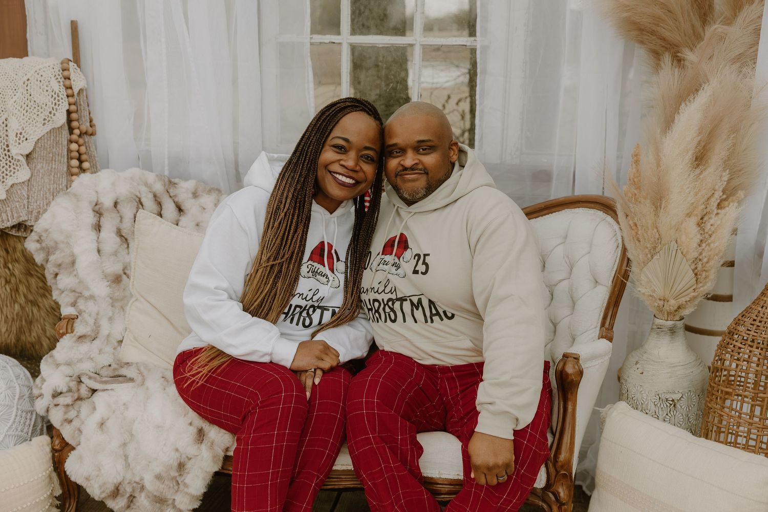 A couple wearing matching cream sweaters and red plaid pajama pants pose together for holiday photos.