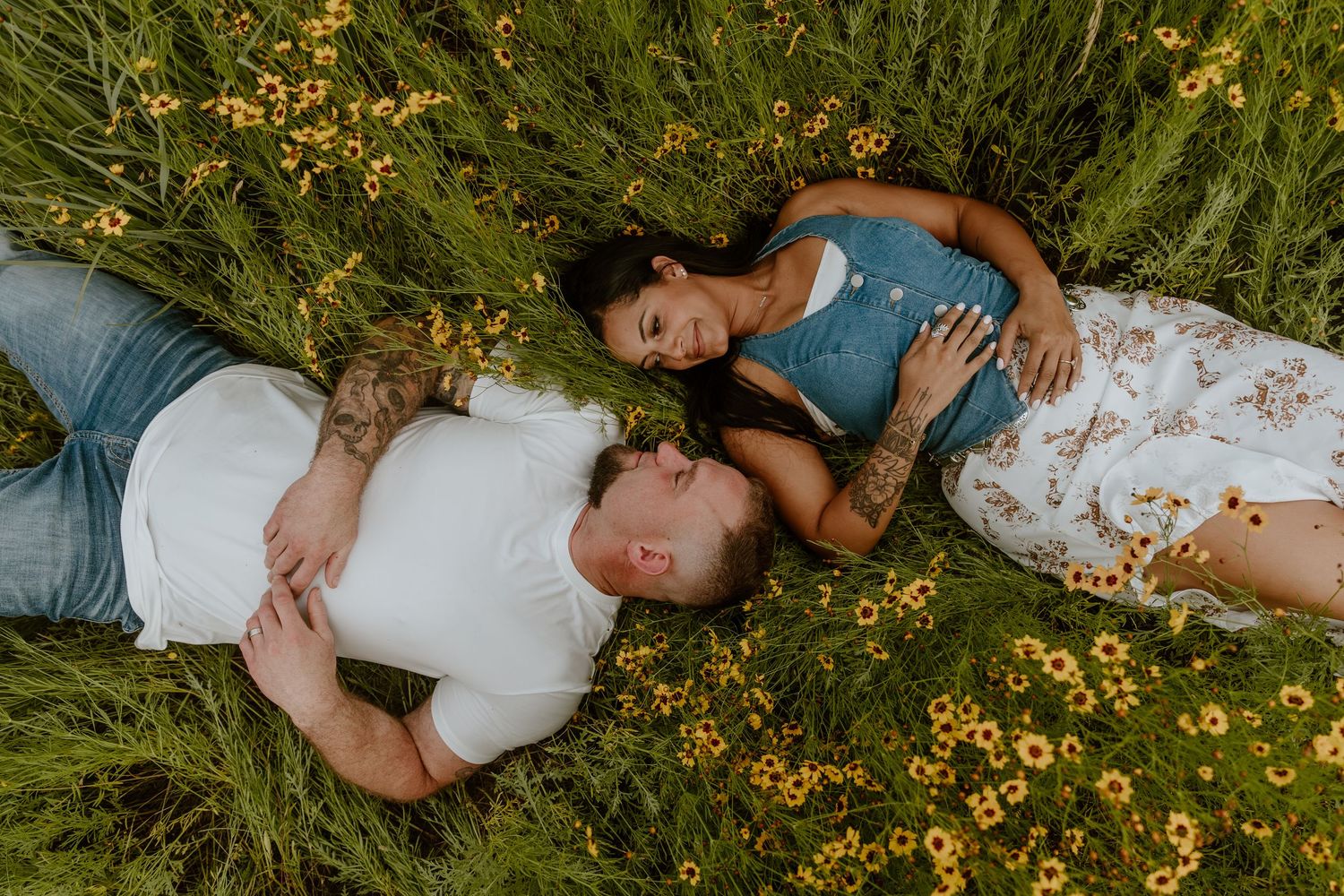 Couple lying in a wildflower field, smiling at each other, surrounded by green grass and yellow blooms.