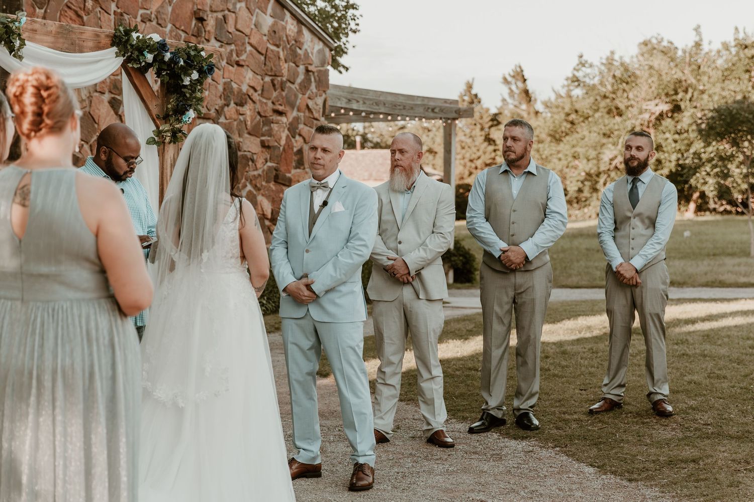 Wedding party in soft blue and gray attire gather outdoors during a rustic summer ceremony.