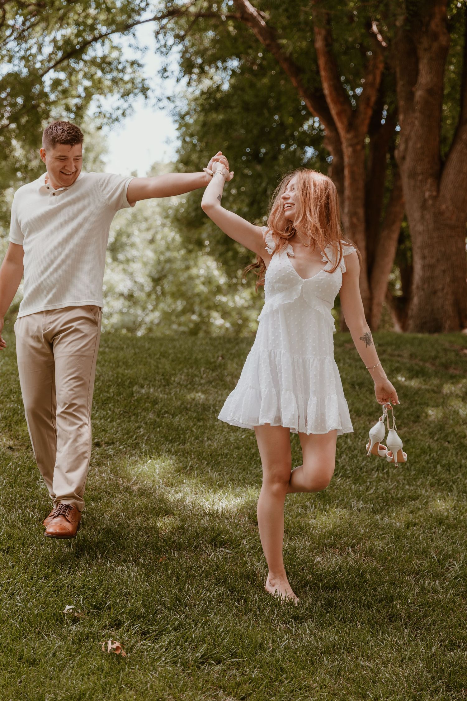 A couple in white clothing dances joyfully together on a grassy hillside under shady trees in a romantic moment.