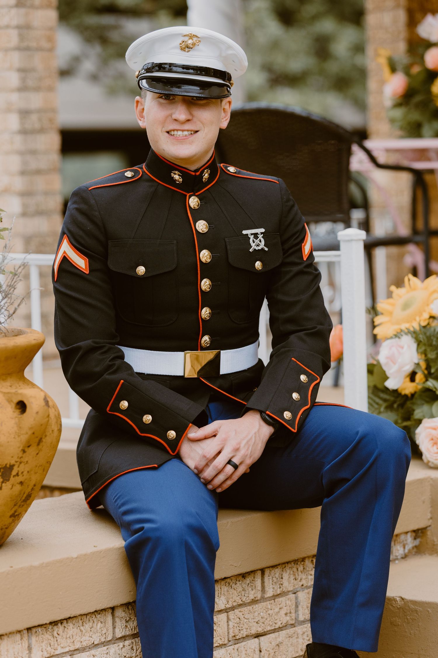 Marine in dress blue uniform sits on stone wall in outdoor setting with decorative planters.