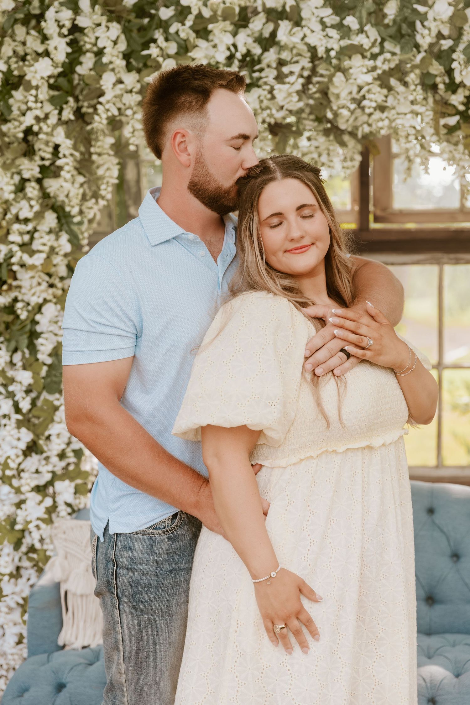 Couple embracing among white floral backdrop, man in blue polo kissing woman in cream dress during engagement photo.