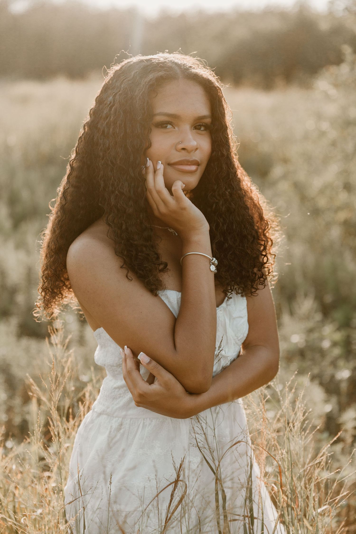 A golden hour portrait of a woman in a white dress with natural curly hair standing in a sun-lit field.