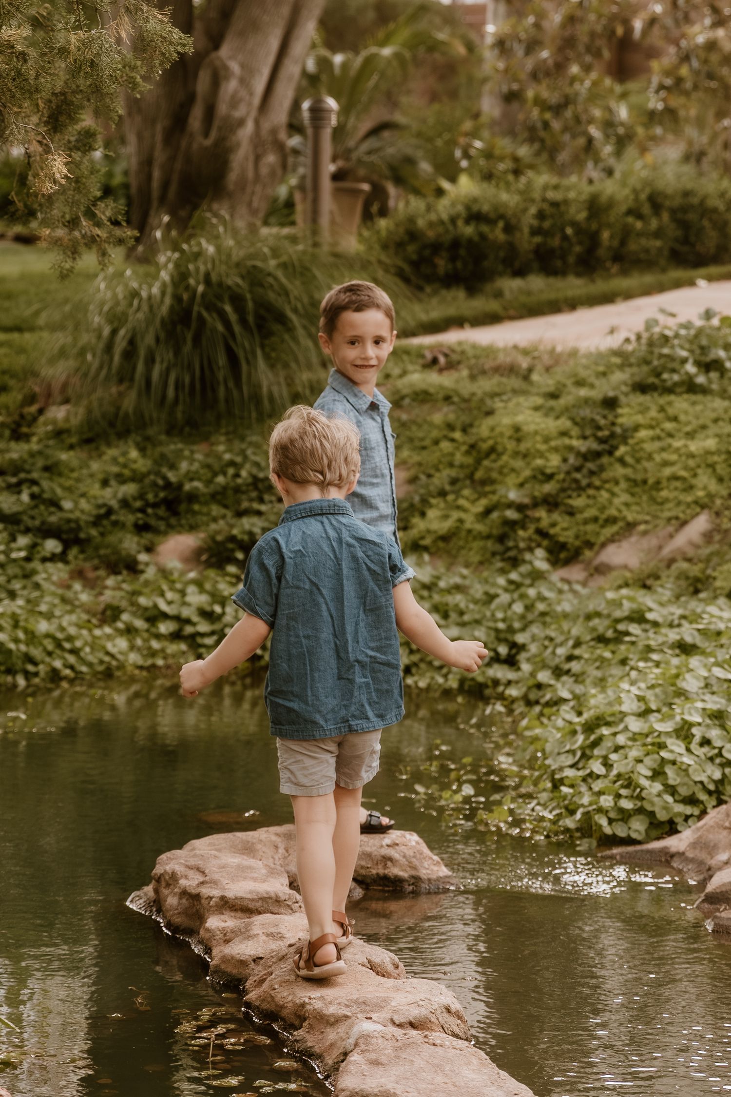 Two young boys in blue shirts balance on rocks by a pond in a lush green park setting.