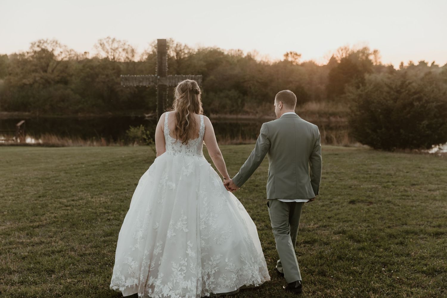 Bride and groom walking hand in hand through a green field at sunset, bride in lace ballgown.