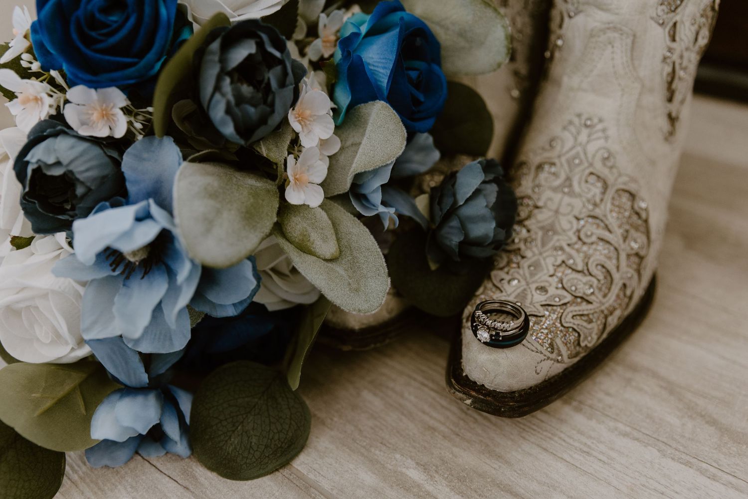 Wedding details featuring a white and blue floral bouquet next to an ornate lace cowboy boot on wooden flooring.