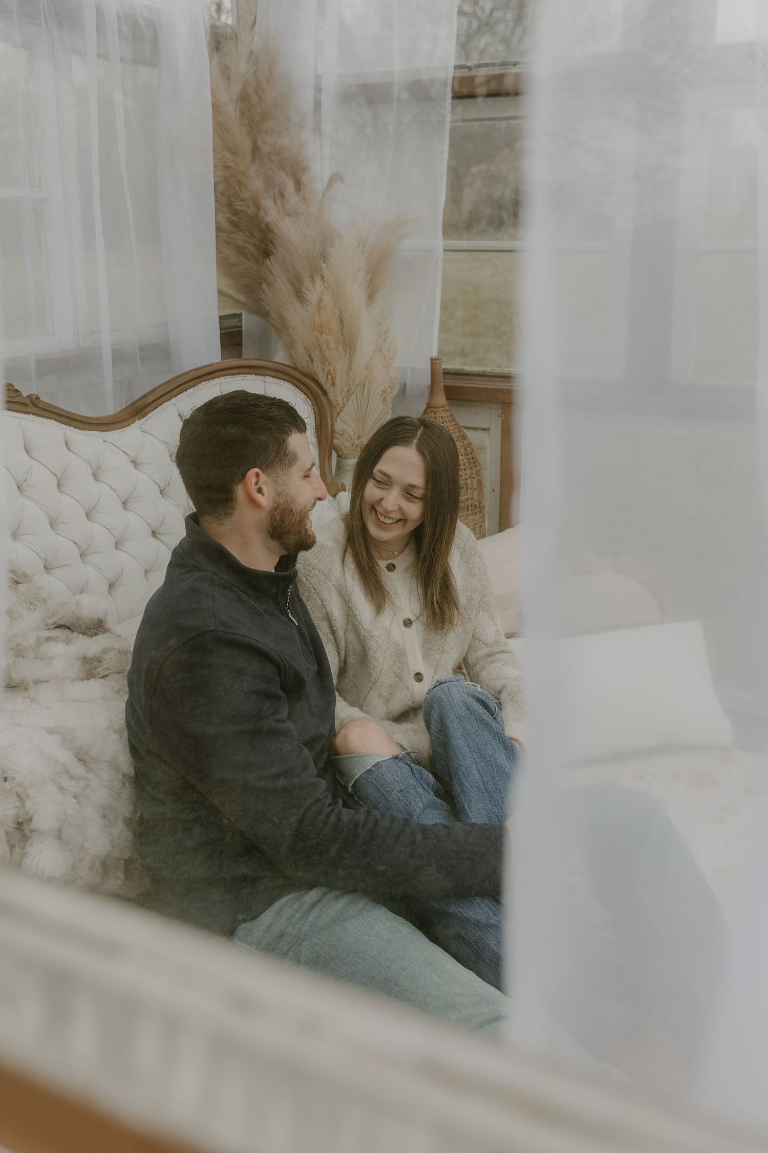 A couple shares an intimate moment while sitting on a white tufted couch near sheer curtains.