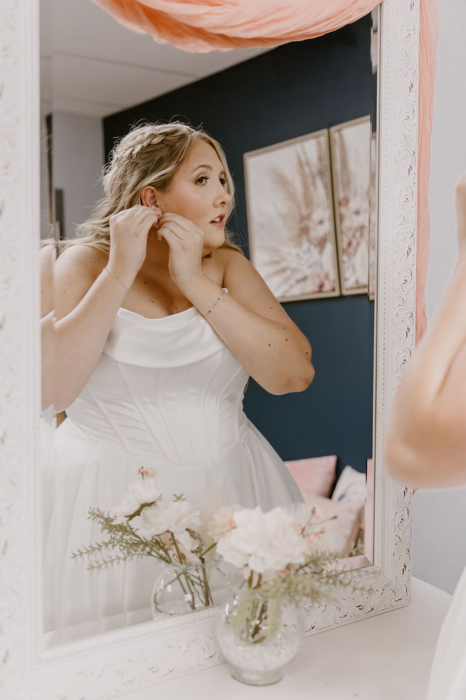 A bride in a white dress adjusts her earring while looking in an ornate mirror with pink balloons in the background.
