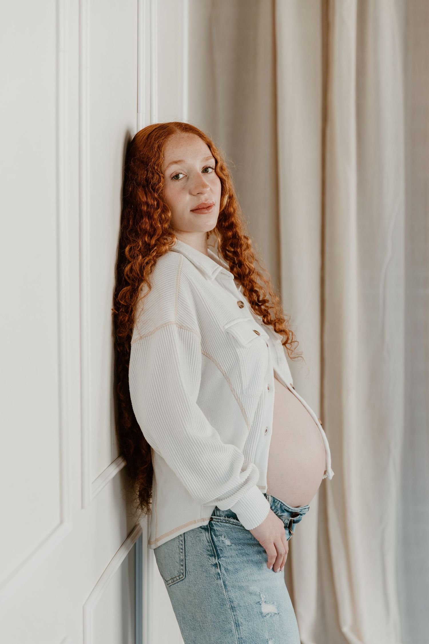 A person with curly red hair wearing a white blazer and light jeans poses against a white curtain background.