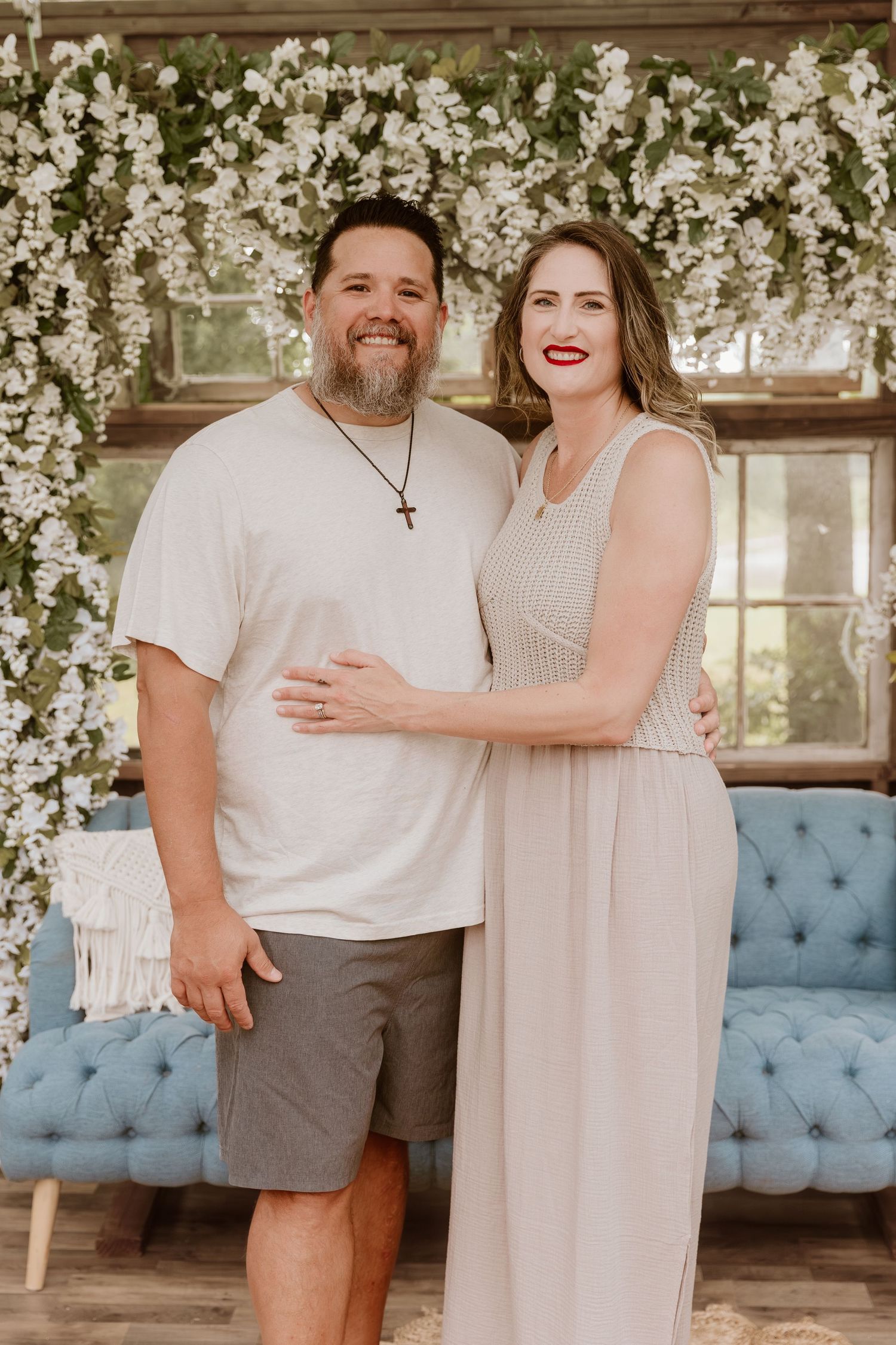 A smiling couple poses together in front of a floral backdrop with white blooms and a blue tufted sofa.