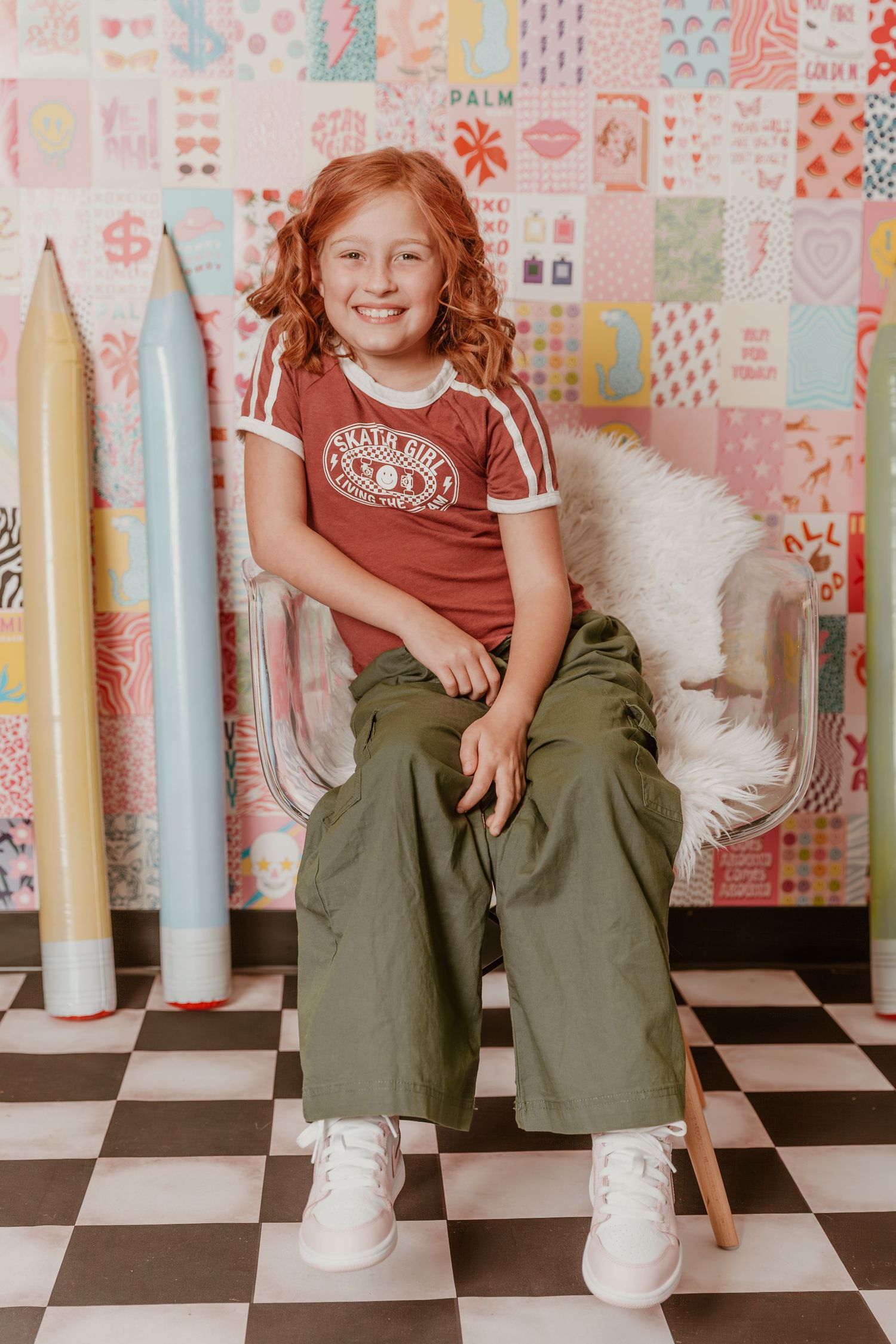 A child sits on a chair wearing a retro style t-shirt and wide leg pants against a patterned wall with surfboards.