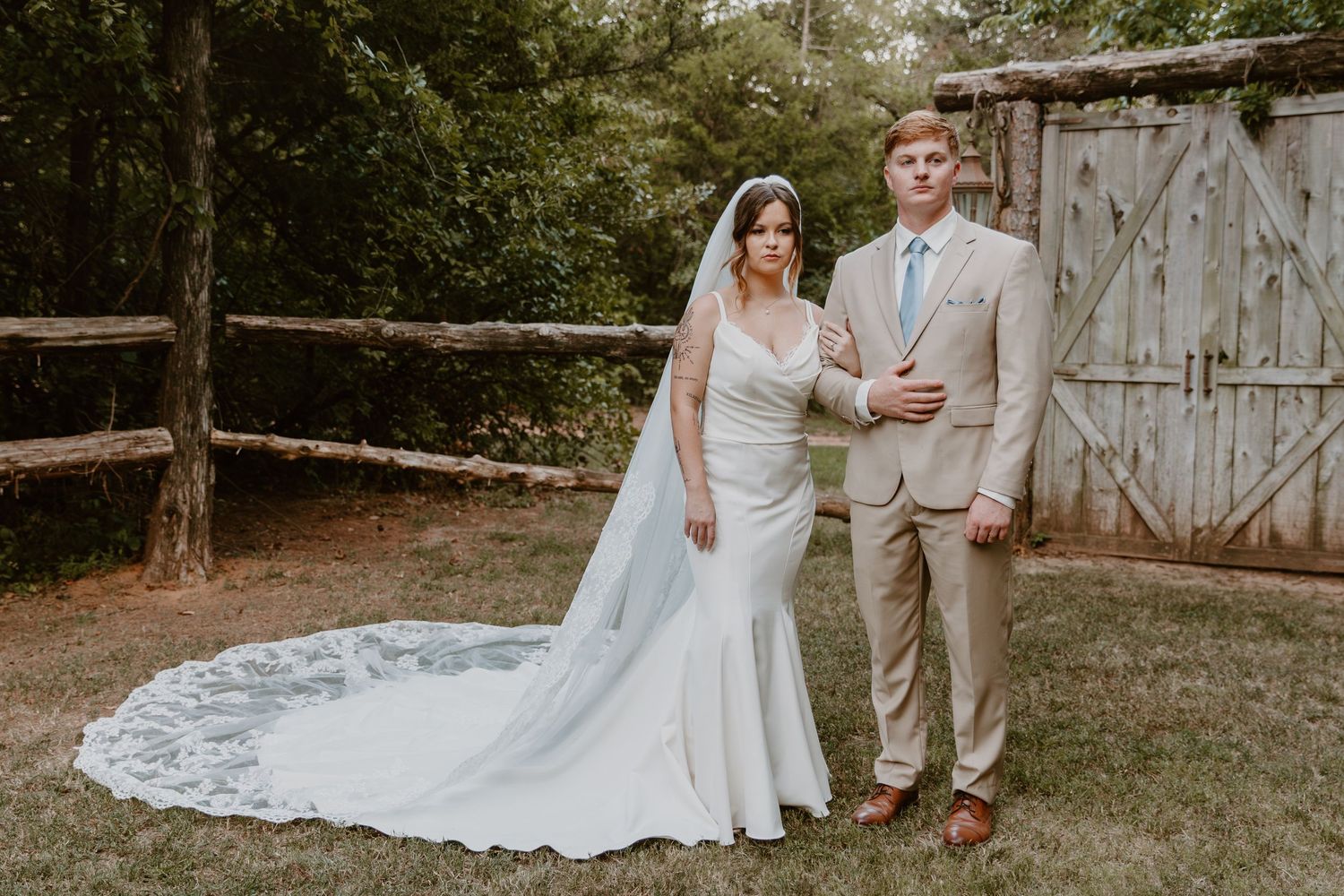 Stylish wedding couple poses in front of rustic barn doors with the bride's lace veil gracefully flowing behind them.