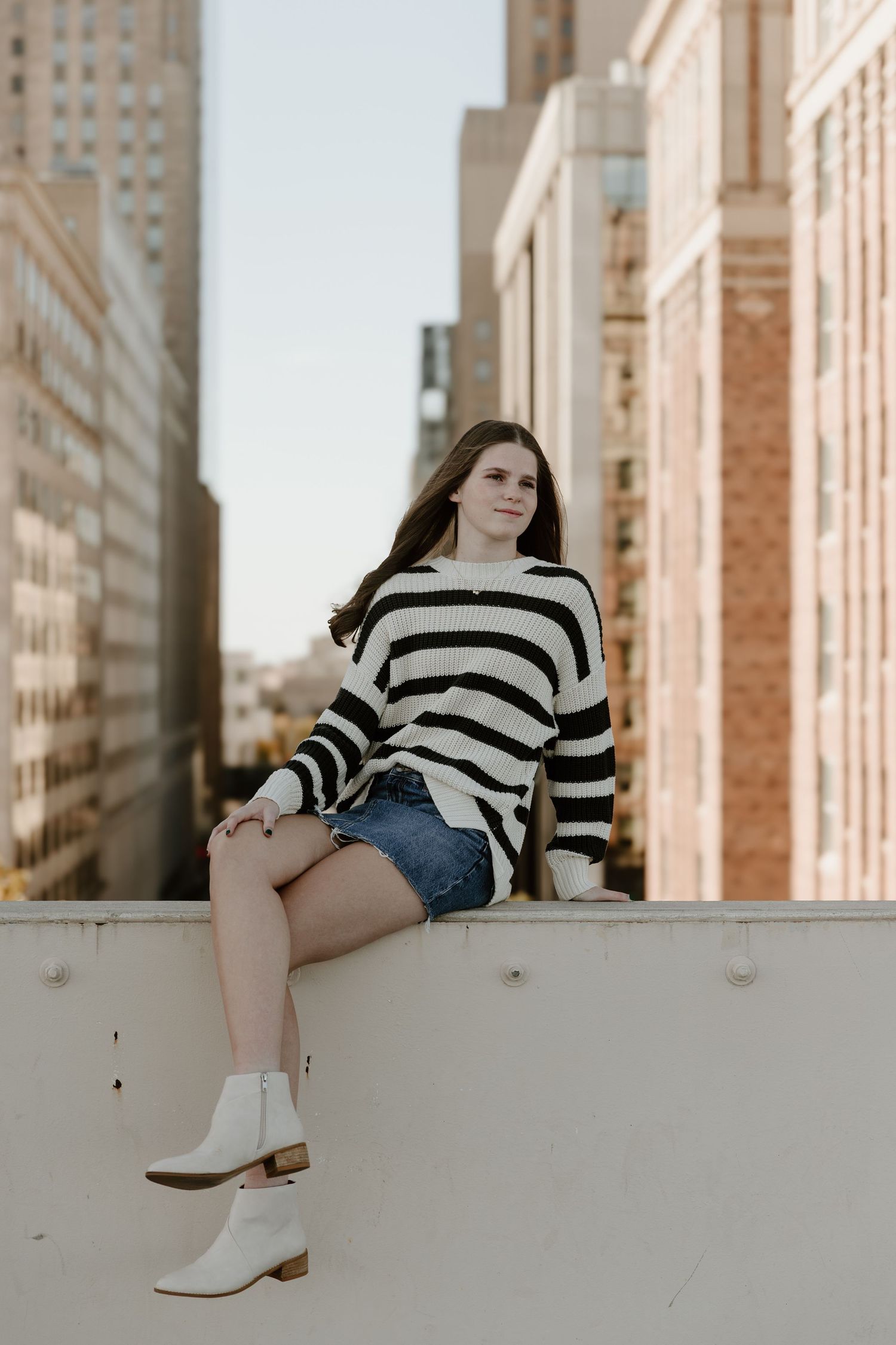 Woman in striped sweater and denim shorts sitting on white ledge in urban setting with city buildings in background.