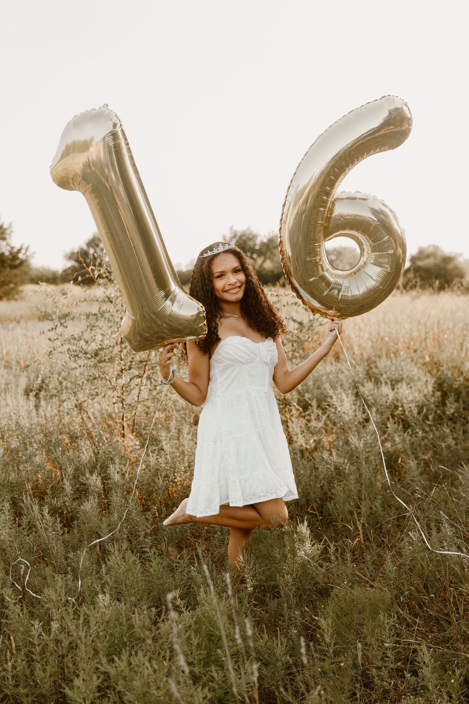 A celebratory sweet 16 photo with gold number balloons in a field at sunset.