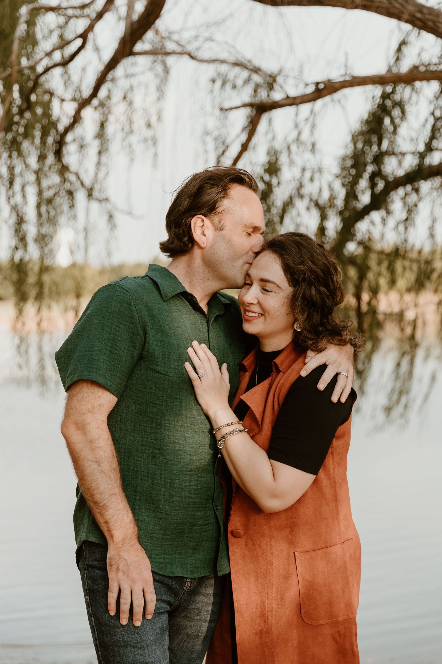 A couple shares tender moments by a lake at sunset, wearing green and rust-colored clothing.