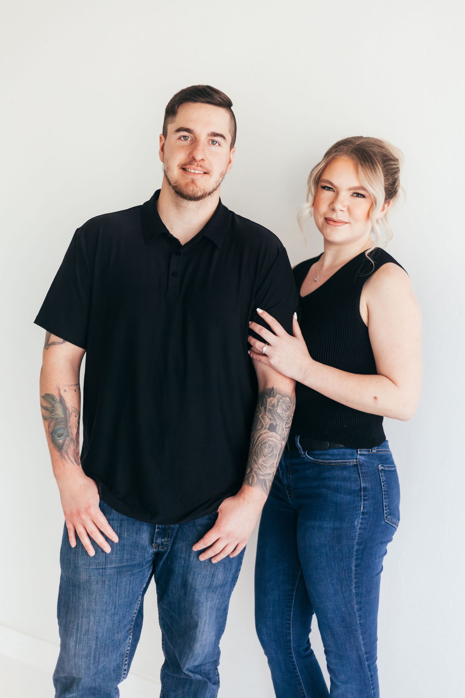 Couple in casual attire posing against white wall for engagement photos.