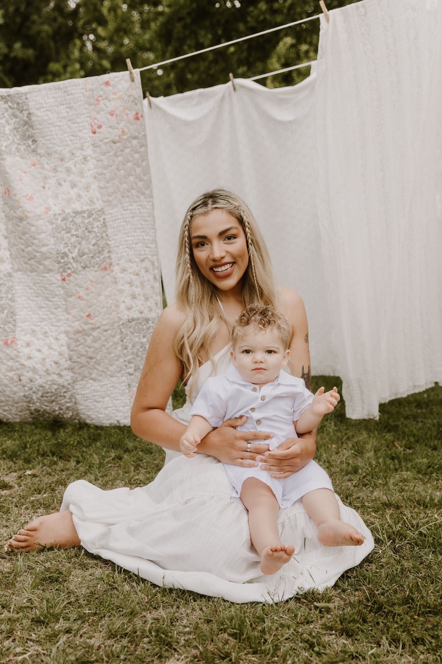 A smiling blonde woman in a white dress sits on grass holding a baby in white outfit, with laundry hanging on a clothesline behind them.