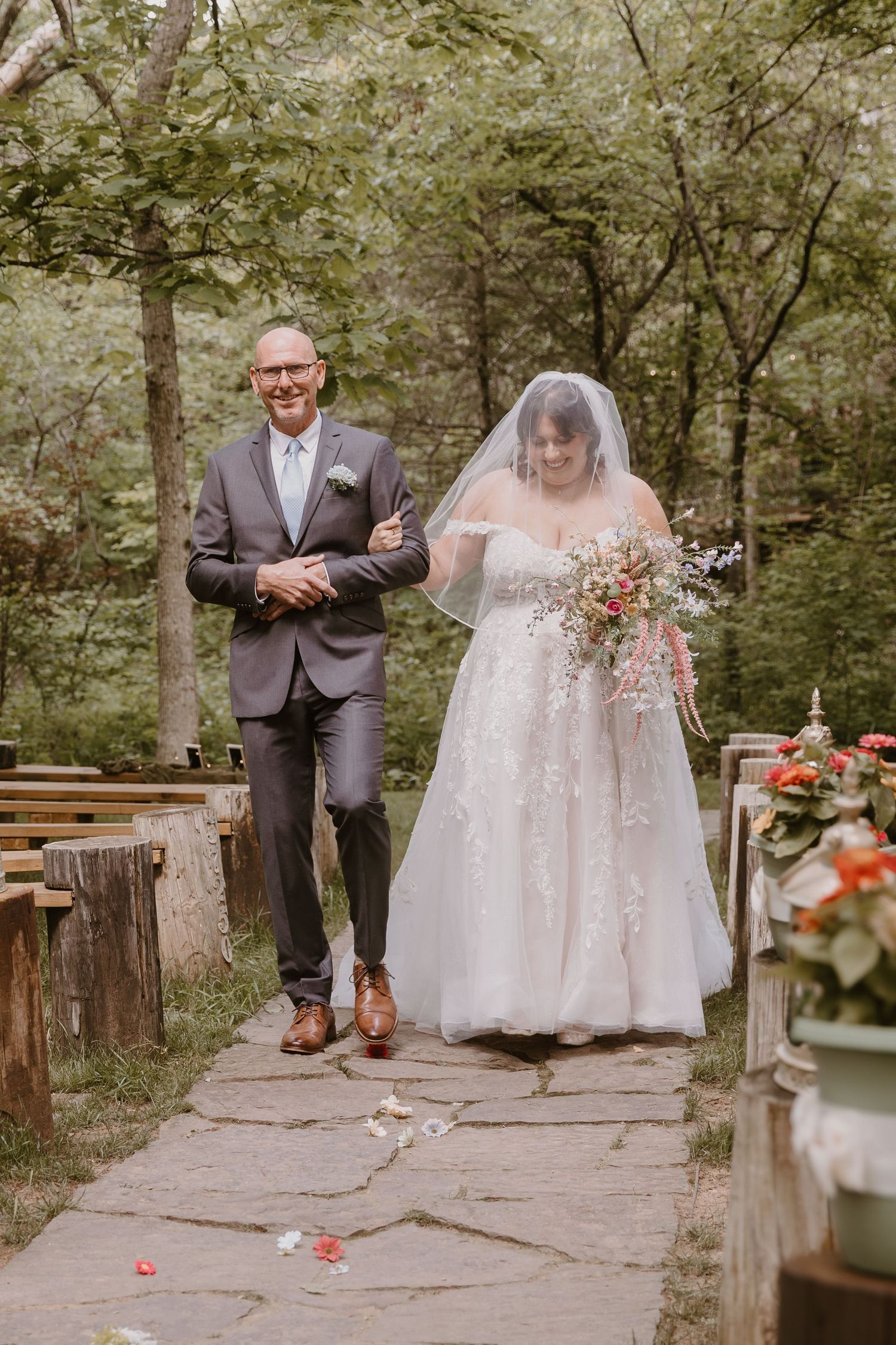 Bride and father walk down outdoor stone aisle surrounded by trees at a woodland wedding ceremony.