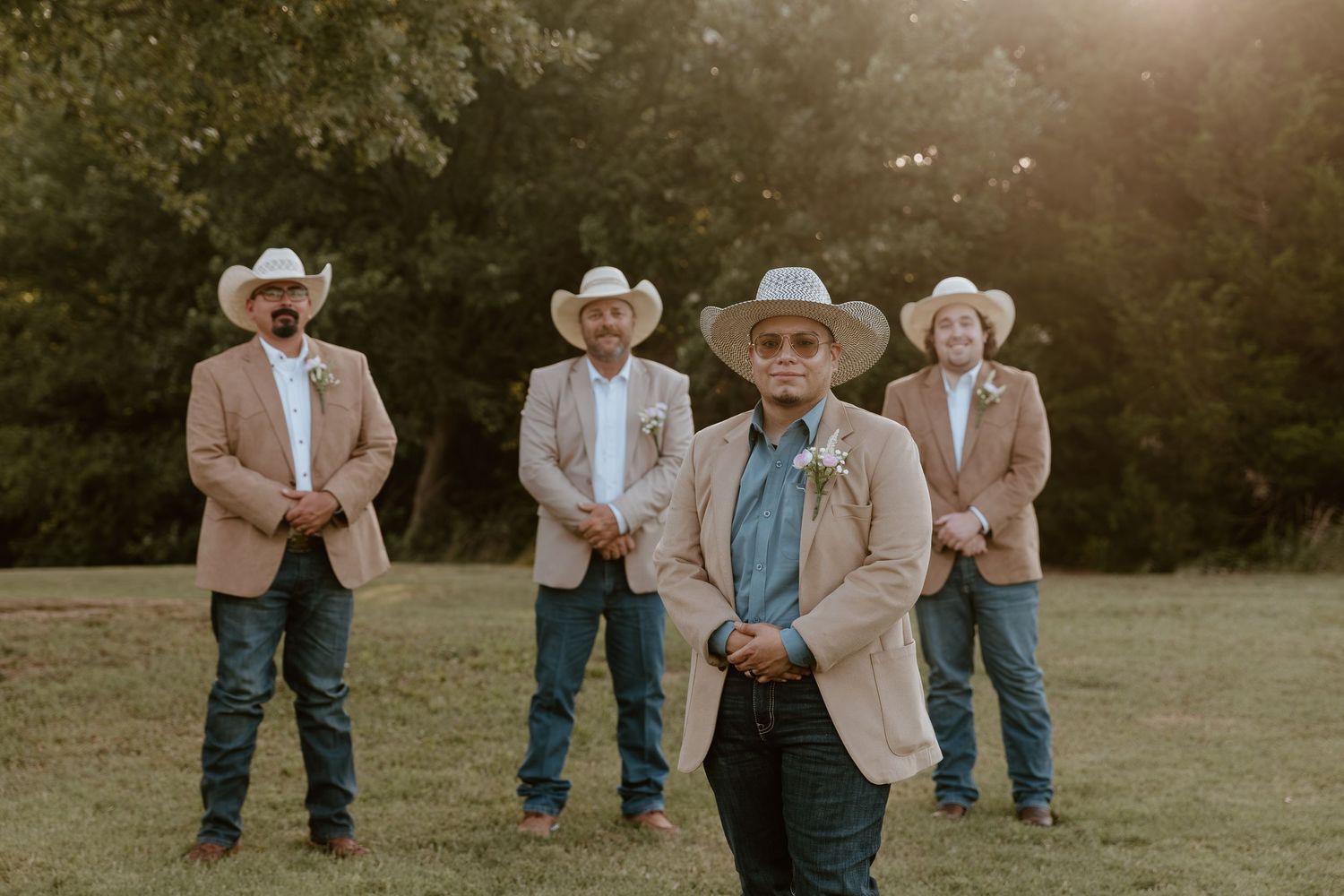 Four groomsmen in cowboy hats and tan blazers with jeans pose outdoors at a rustic country wedding.