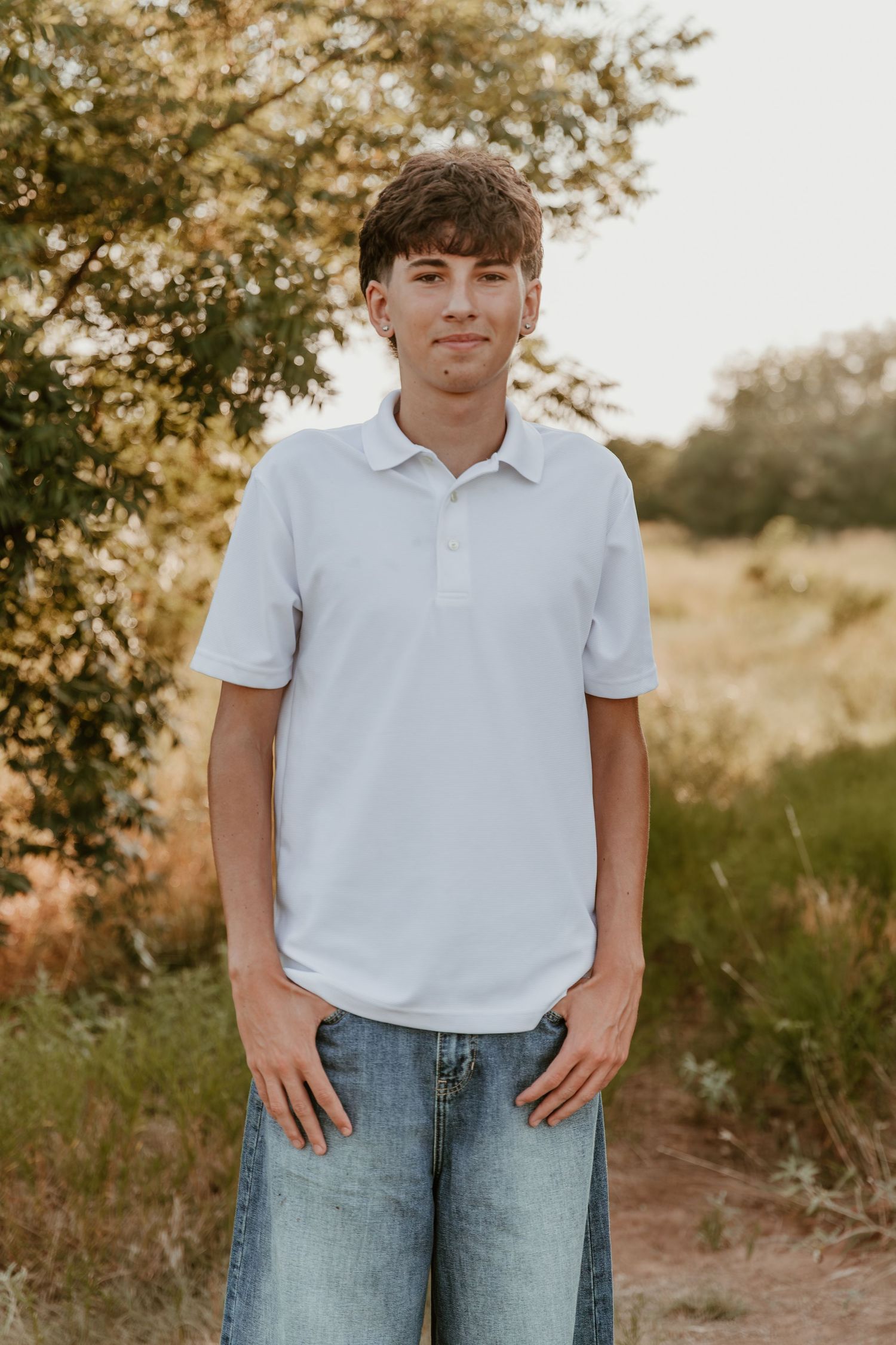 A figure in a white polo shirt and jeans stands outdoors against a natural background with trees at sunset.