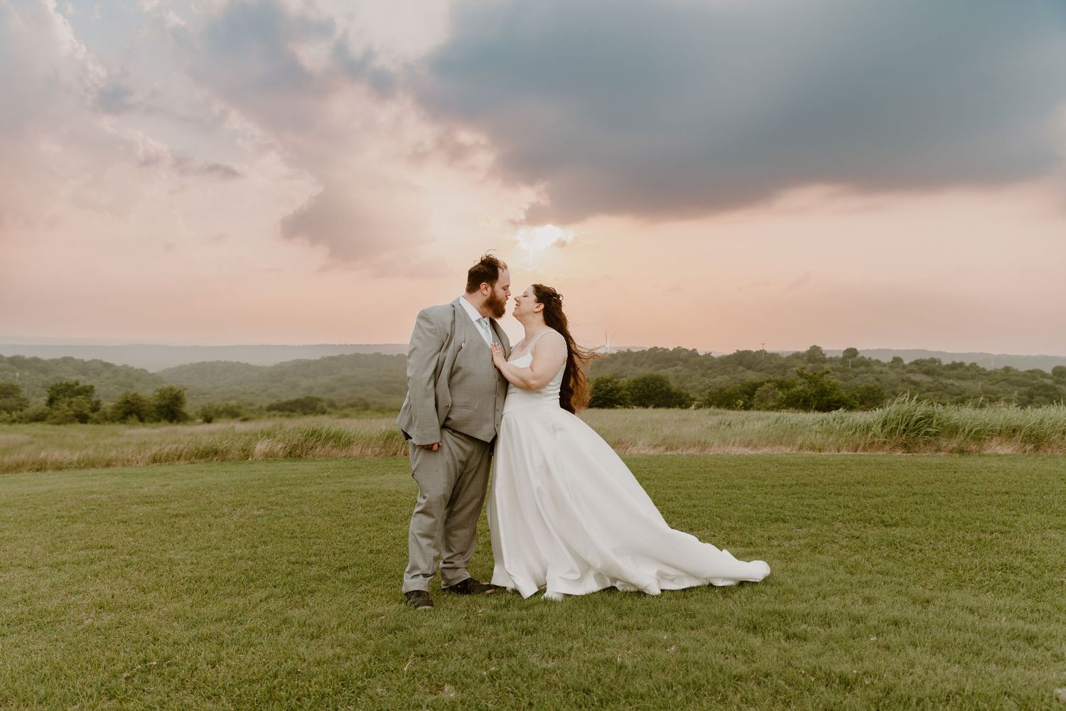 Romantic newlyweds walk hand in hand across rolling green hills at sunset after their wedding ceremony.