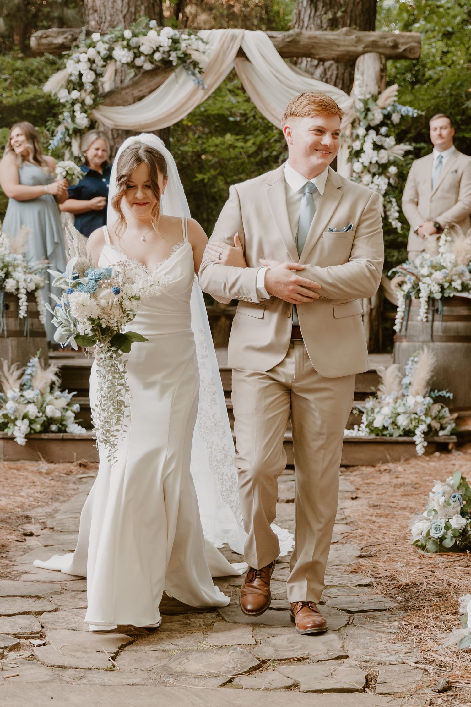 Elegant outdoor wedding ceremony with tan suits and white floral decorations on a stone path surrounded by greenery.