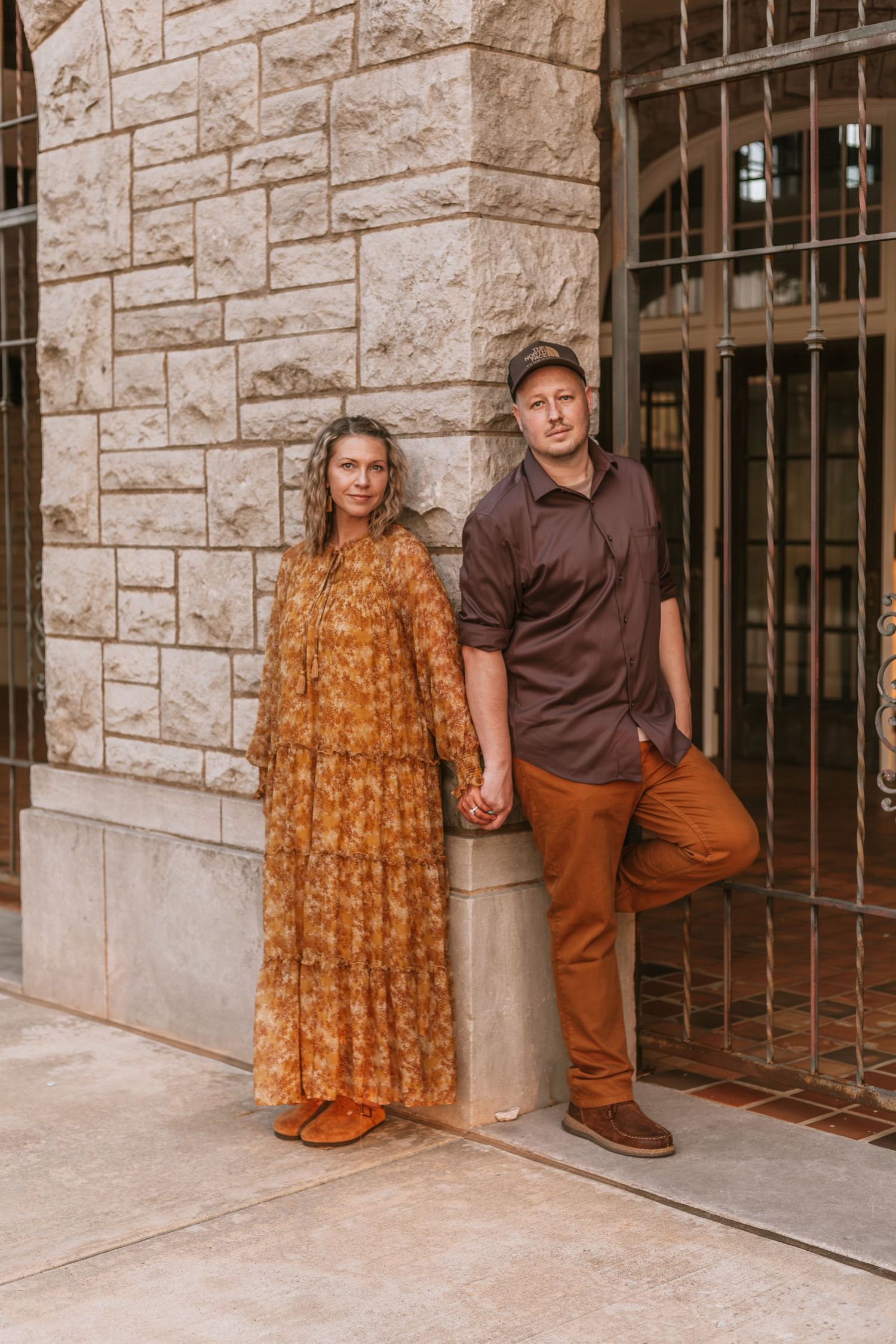 Couple in brown and orange outfits posing against stone wall with wrought iron gates.