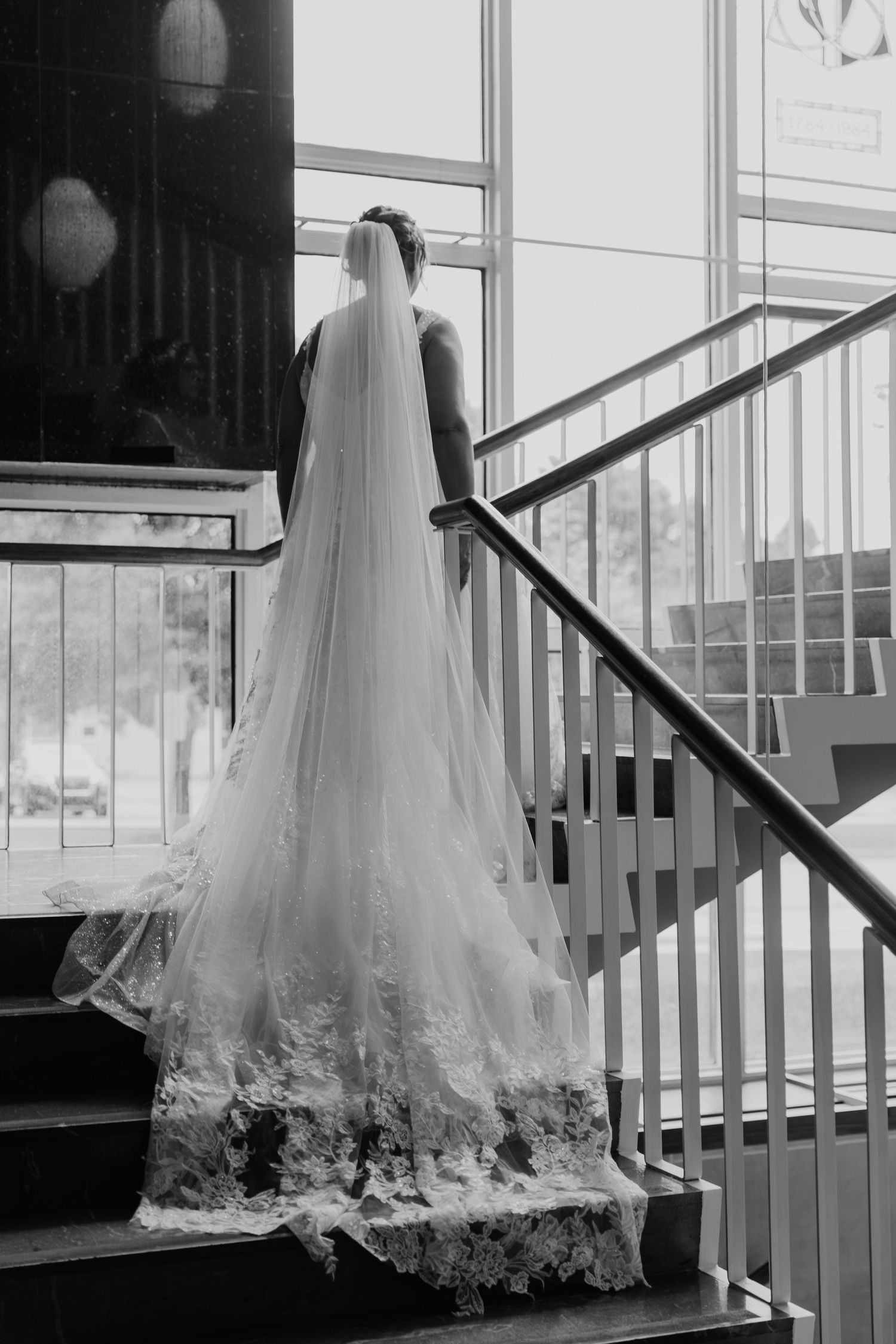 A bride in an elegant white gown with a flowing lace-trimmed veil ascends a modern staircase in black and white.