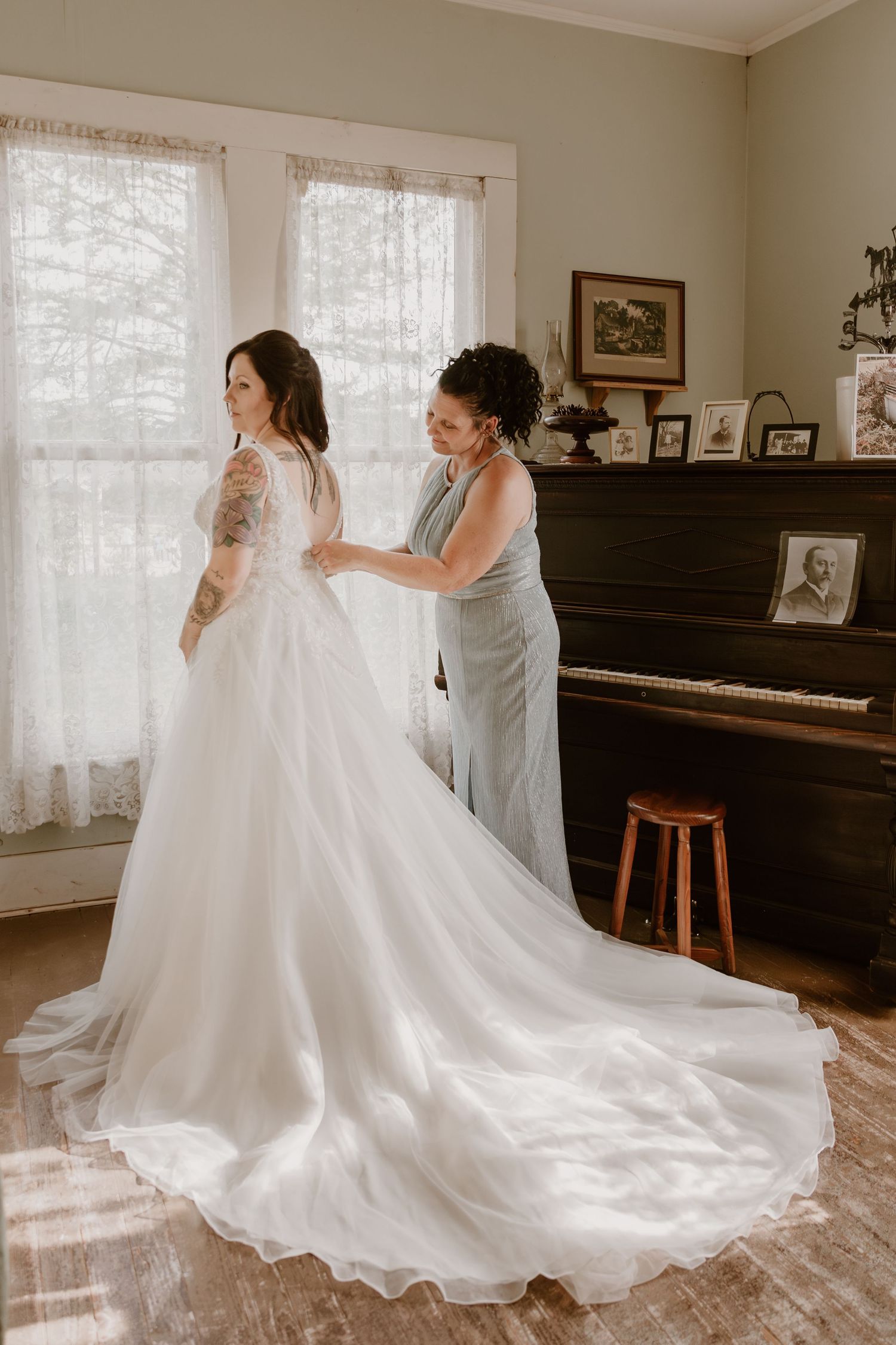 A bride in a flowing white gown with a long train gets help with her dress in a room with a piano and lace curtains.