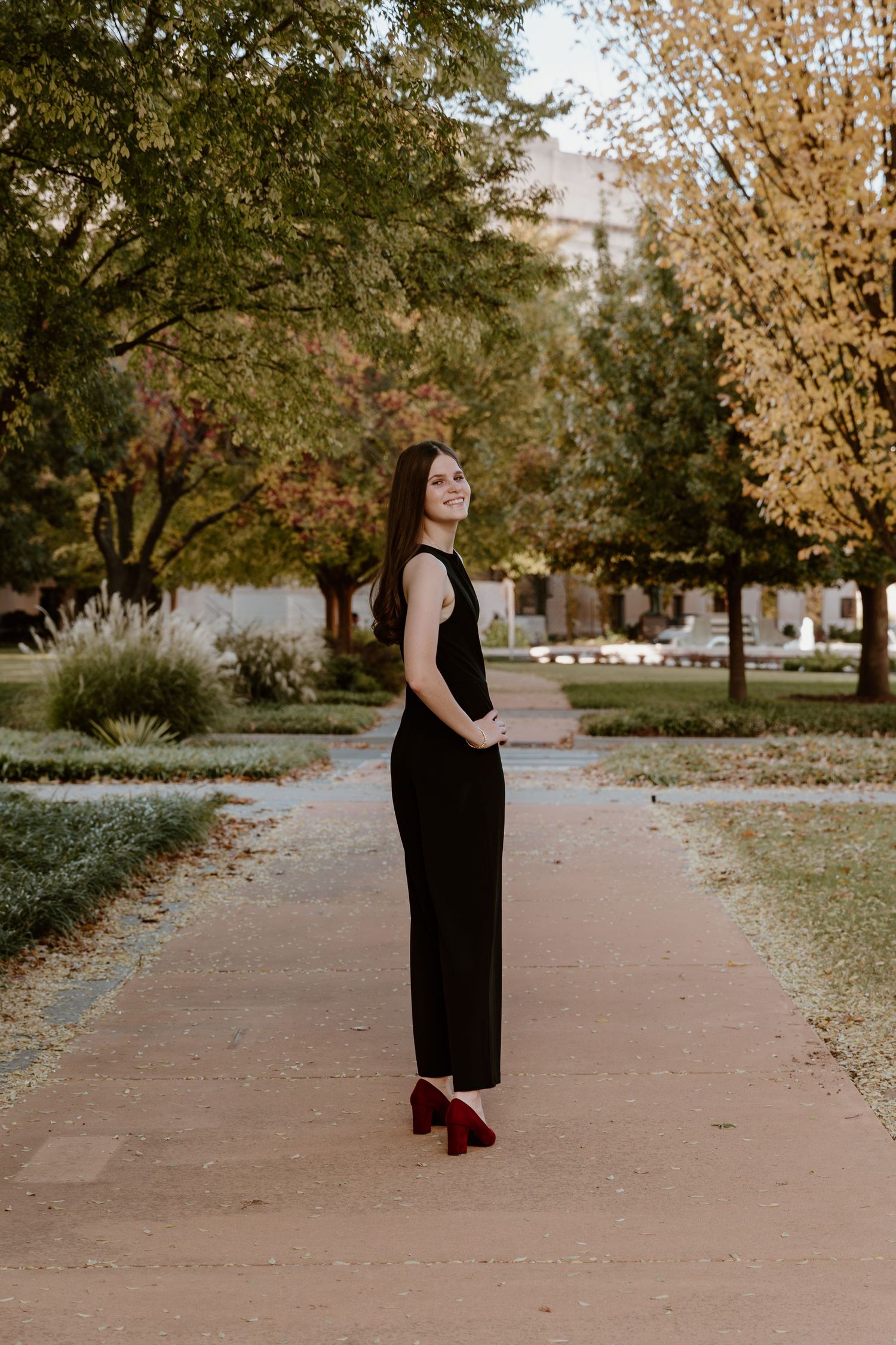 A person in a black jumpsuit and burgundy shoes stands on a path surrounded by autumn trees in a park setting.
