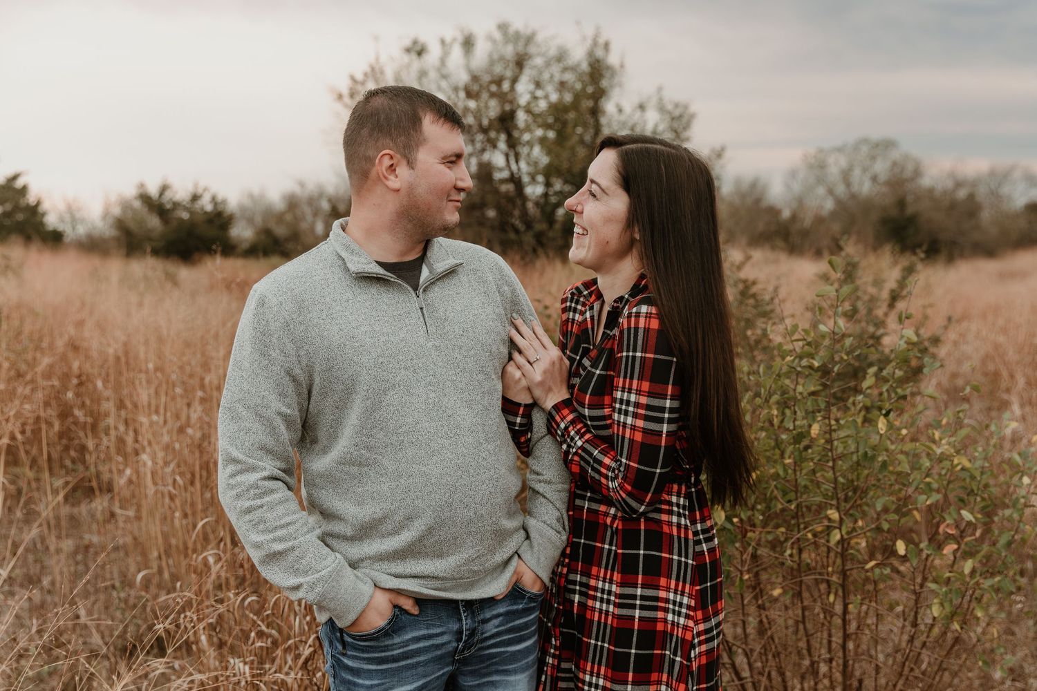 A romantic outdoor photoshoot features a couple in matching plaid and grey sweaters against a rustic field backdrop.
