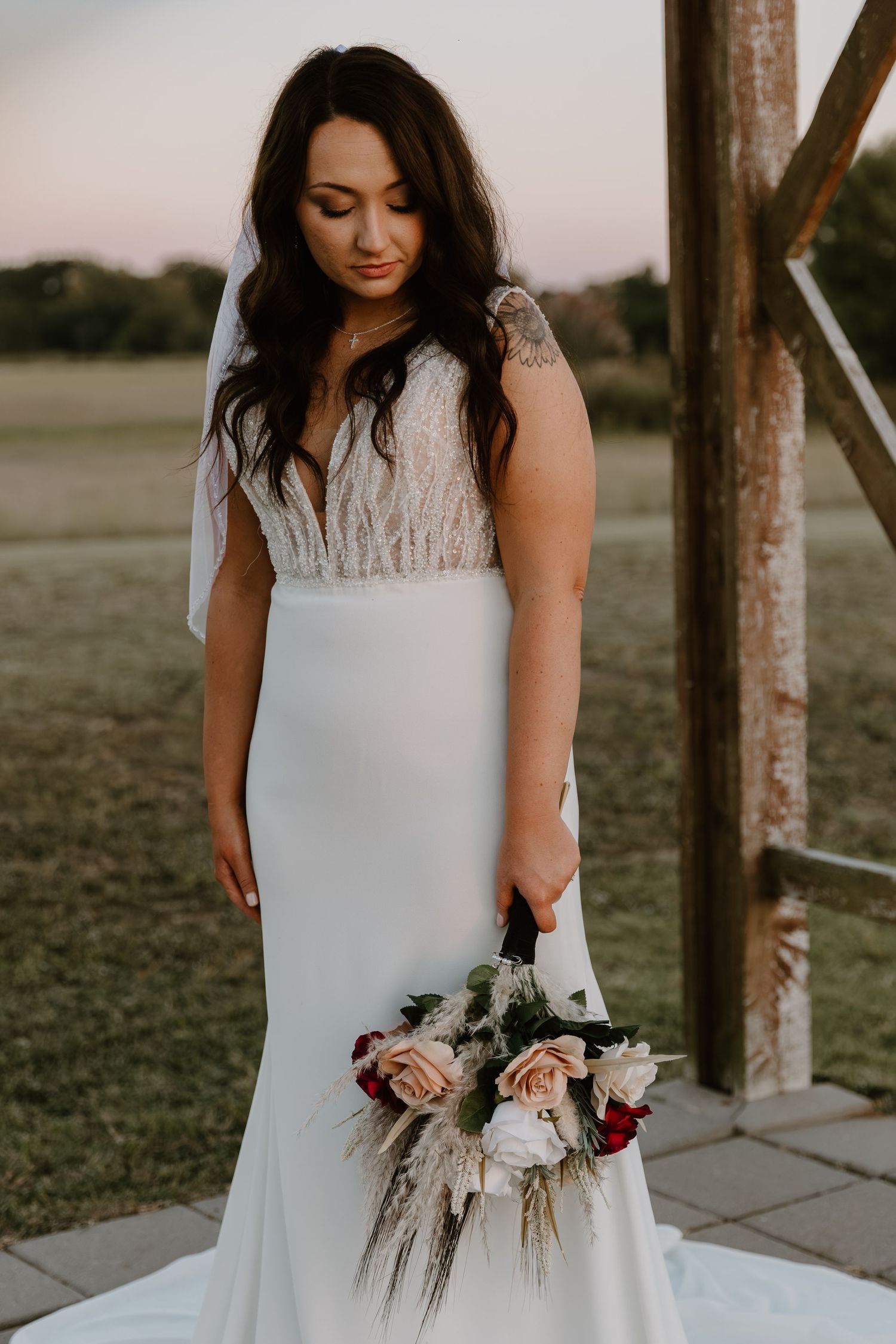 A bride in a fitted white wedding dress with lace bodice holds a romantic bouquet of roses and dried pampas grass at sunset.