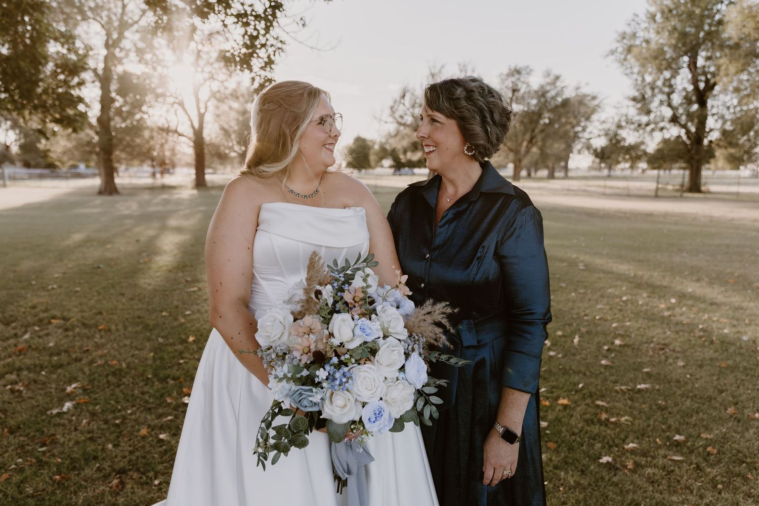 A couple in wedding attire stands together in a sun-drenched autumn field with trees in the background.